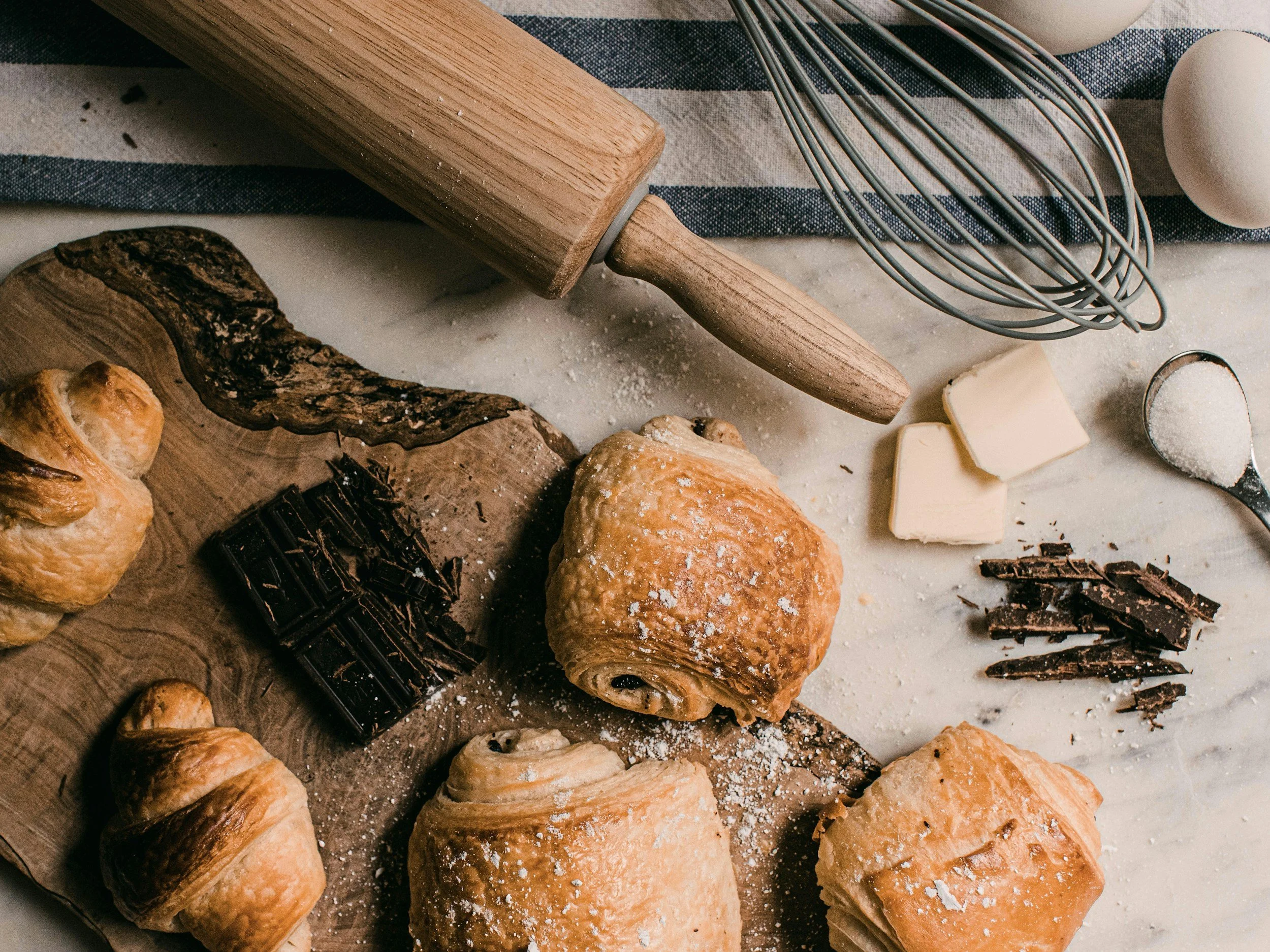 Assorted baked pastries, a rolling pin, a whisk, and a block of white chocolate on a baking surface with flour and chocolate shavings.