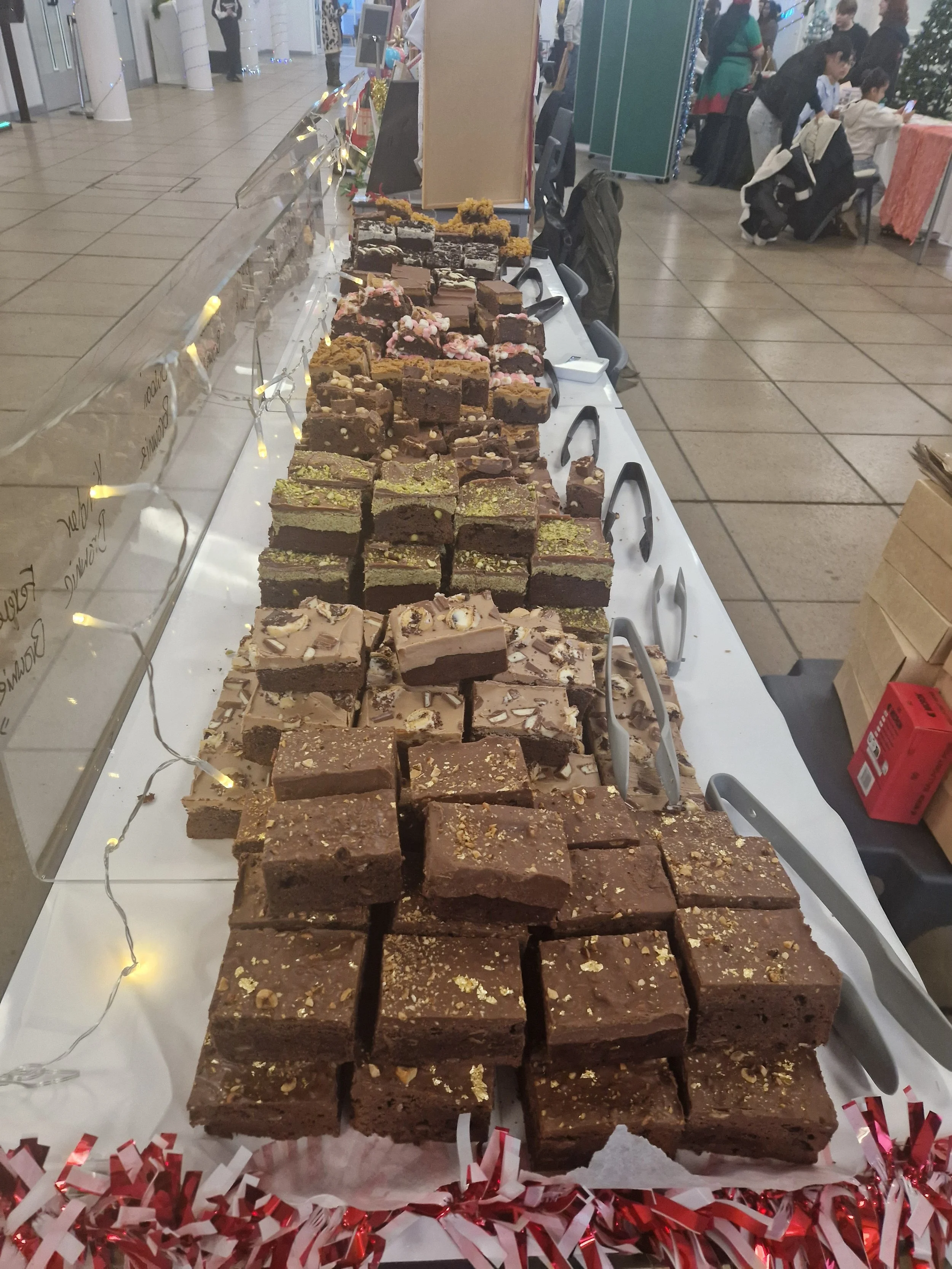 A table filled with various types of brownies at a holiday market, decorated with red and white tinsel.