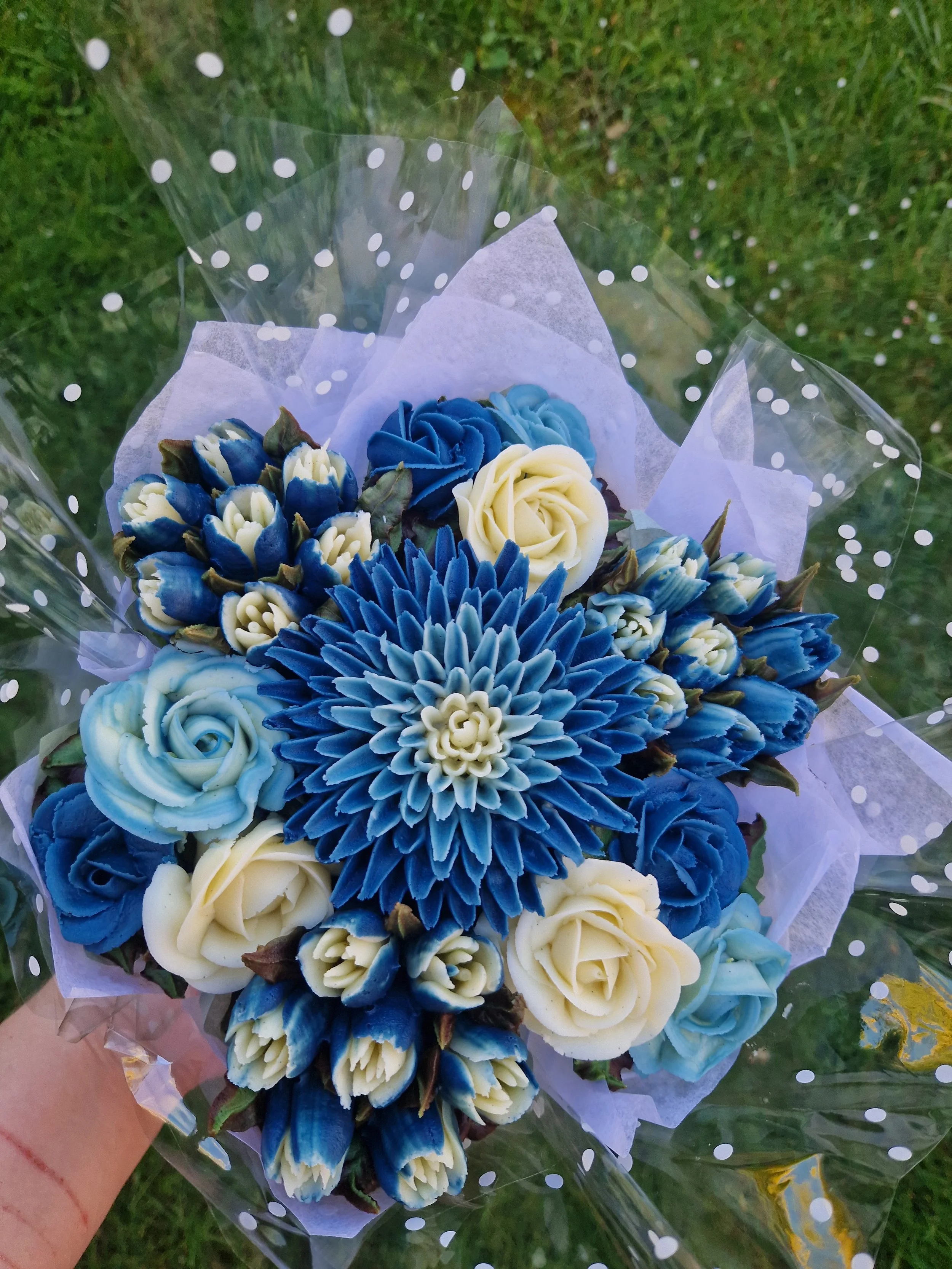 A bouquet of blue and white roses and flowers wrapped in clear polka-dotted plastic, held outdoors on grass.