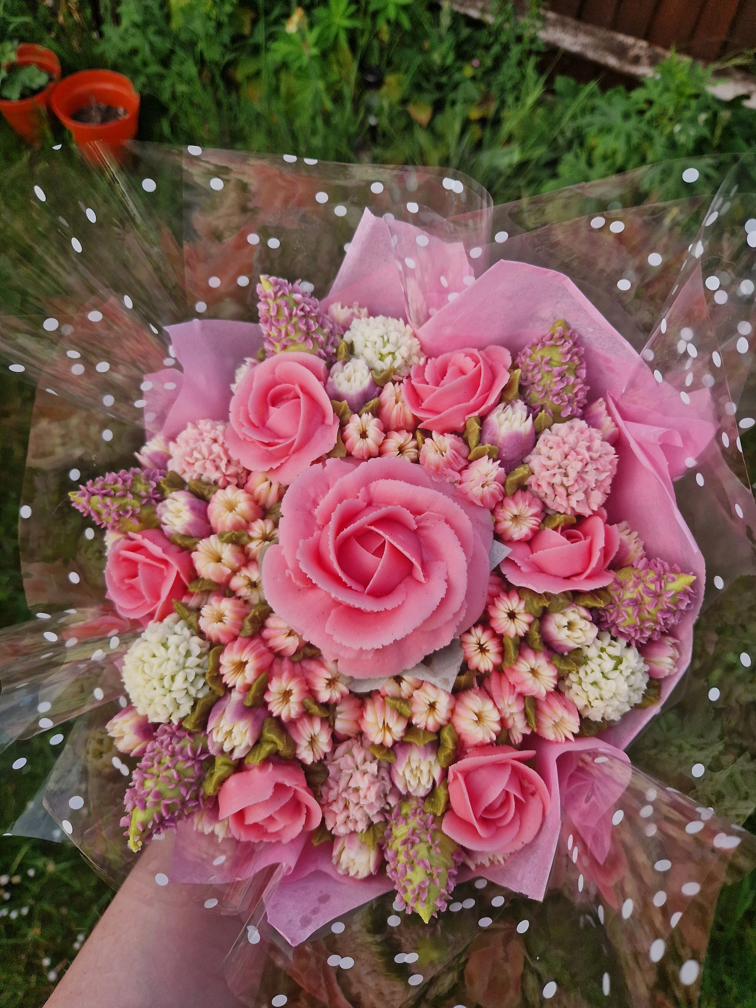 A cake bouquet of pink and white flowers wrapped in pink tissue paper and plastic with white polka dots, held in a person's hand against a garden background.