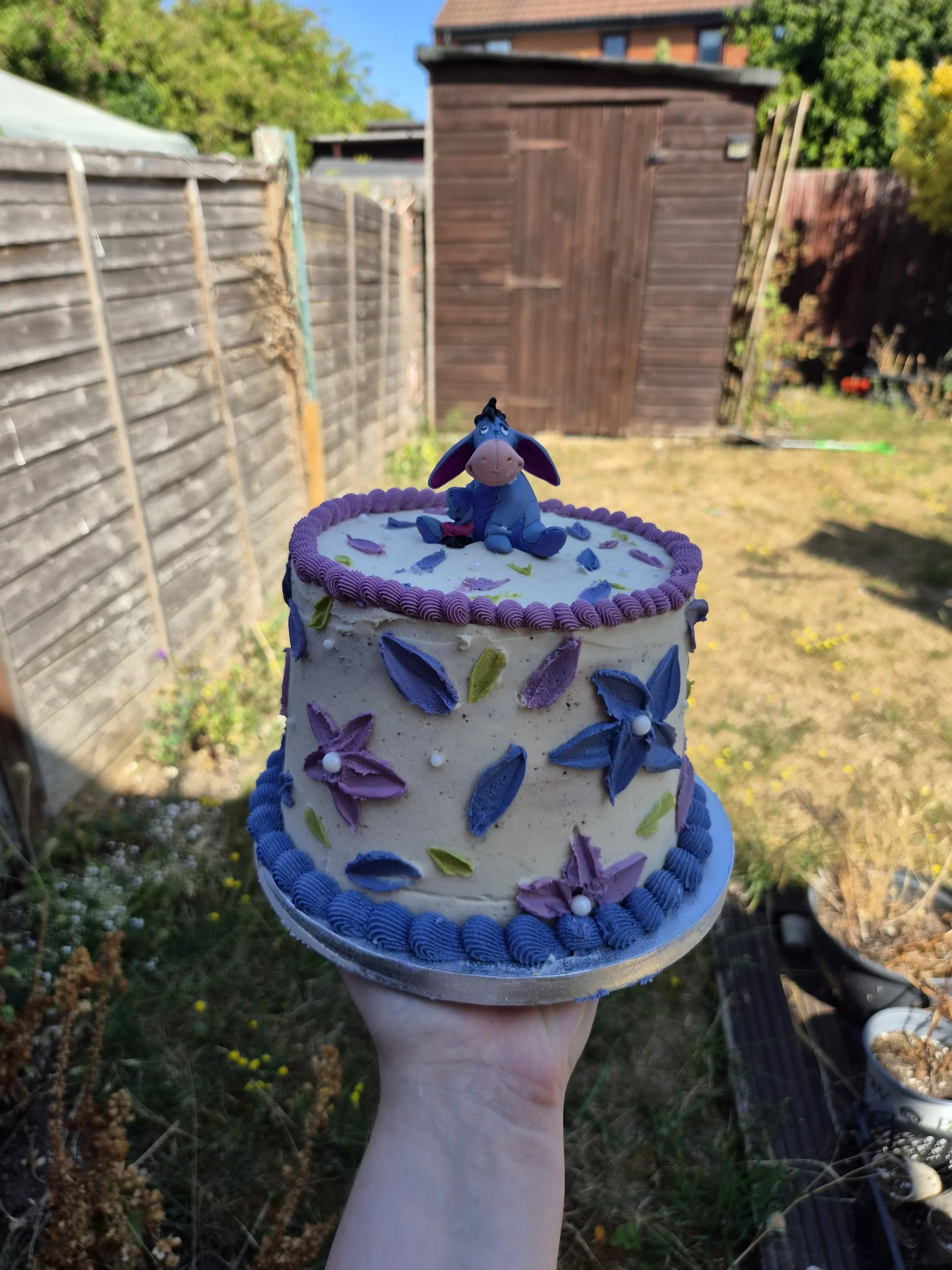 A two-tiered birthday cake decorated with purple, blue, and green flower-shaped icing designs, topped with a small plush Eeyore figurine from Winnie the Pooh, held outdoors in a backyard with fencing, trees, and a wooden shed.