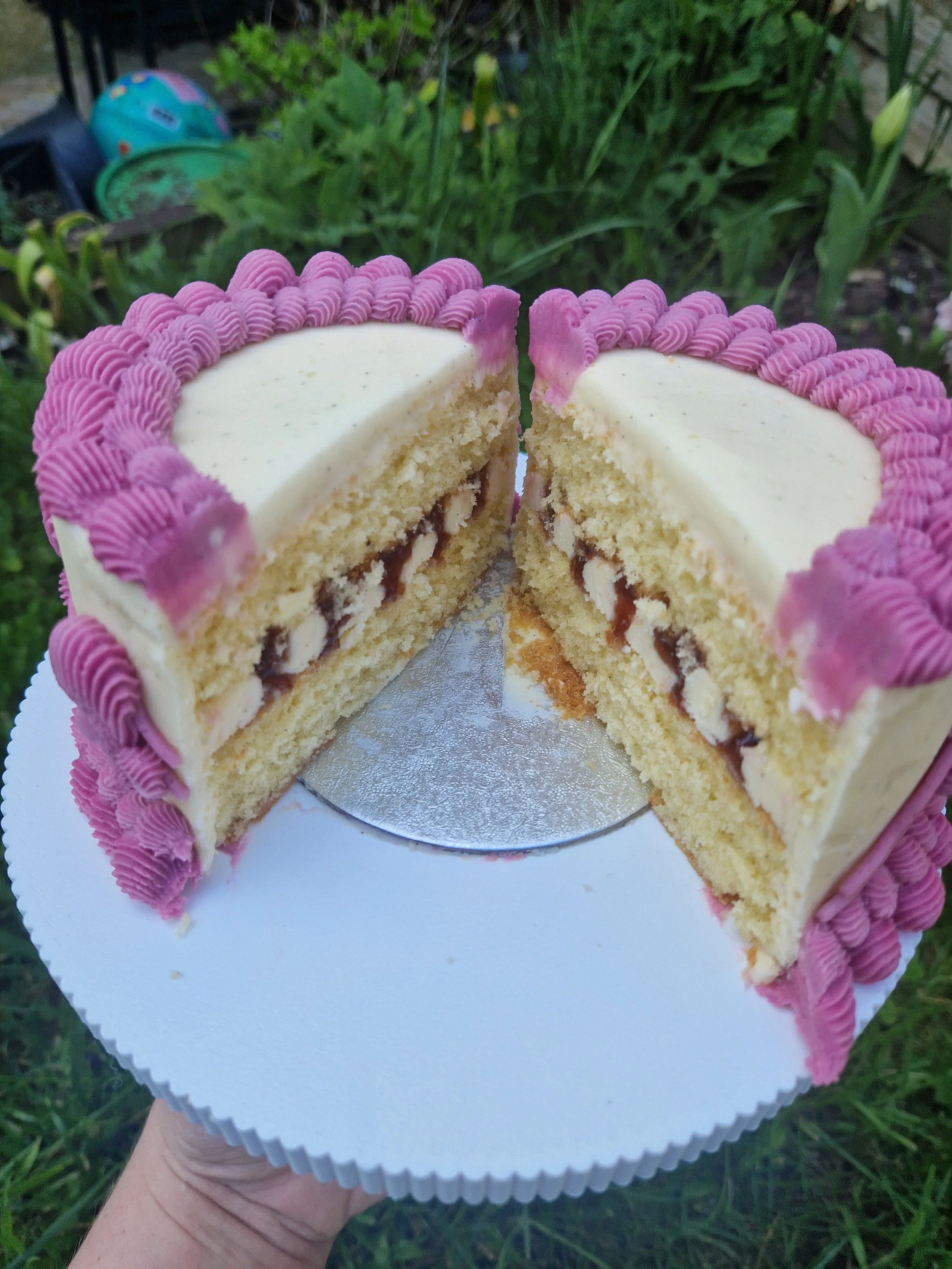 A person holding a partially sliced birthday cake with pink frosting and white icing on top, revealing layers of cake with cream and filling inside. The cake is on a white cake board and is outdoors, with green grass and plants in the background.