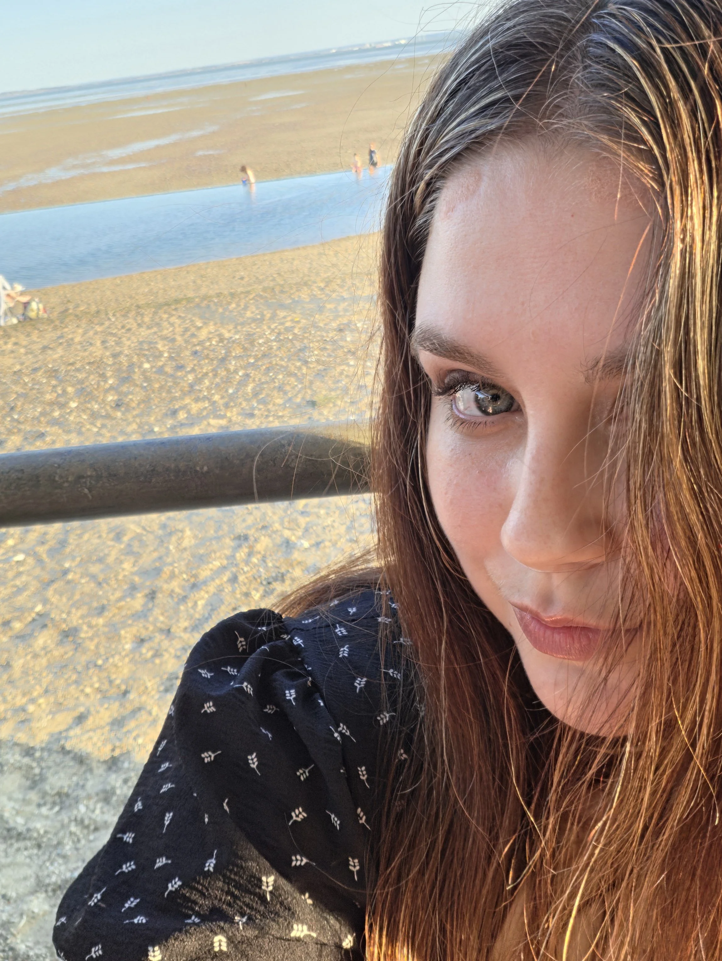 Close-up of a woman's face with brown hair and blue eyes, outdoors at the beach, with the ocean, sandy shore, and a few people in the background.