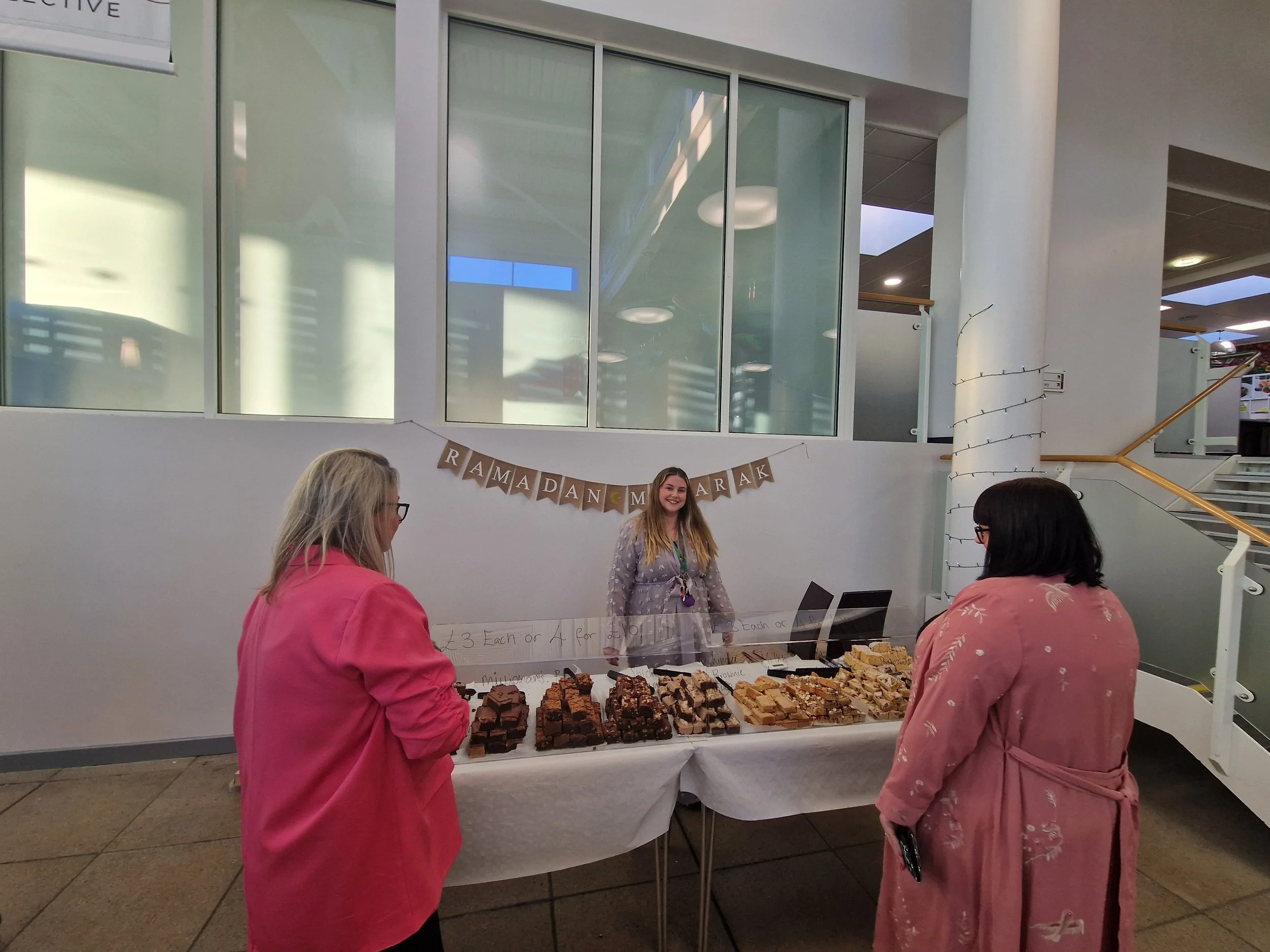 People shopping at a fudge sale table inside a building with large windows and stairs in the background. There is a banner with the word "RAMADAN" visible.