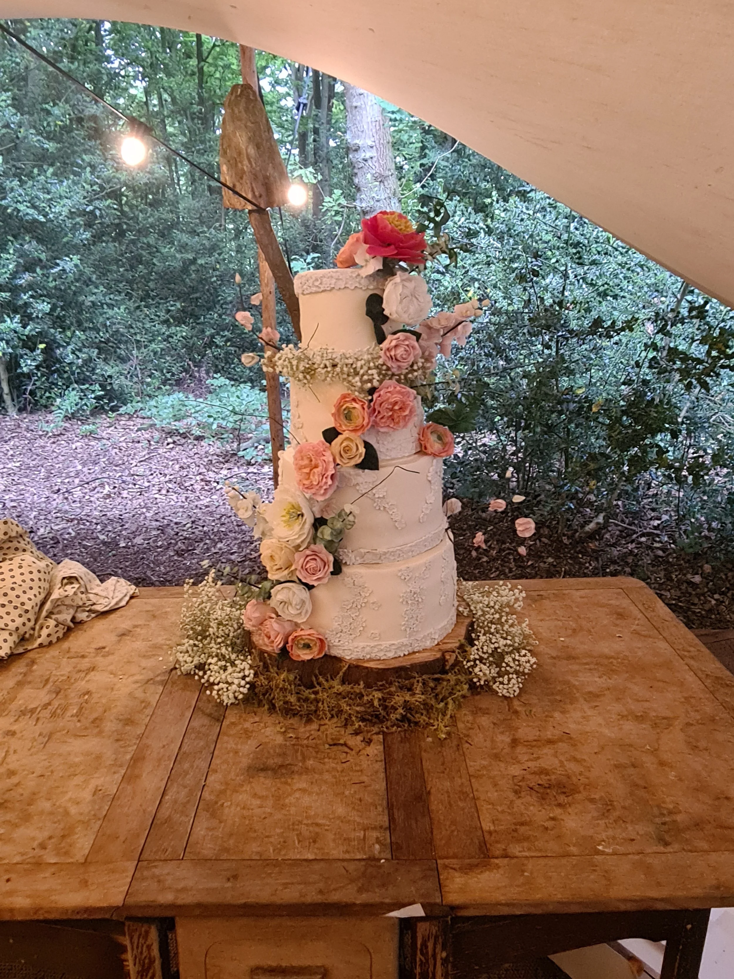 A three-tier white wedding cake decorated with pink and peach roses, baby's breath, and greenery, placed on a rustic wooden table outdoors.