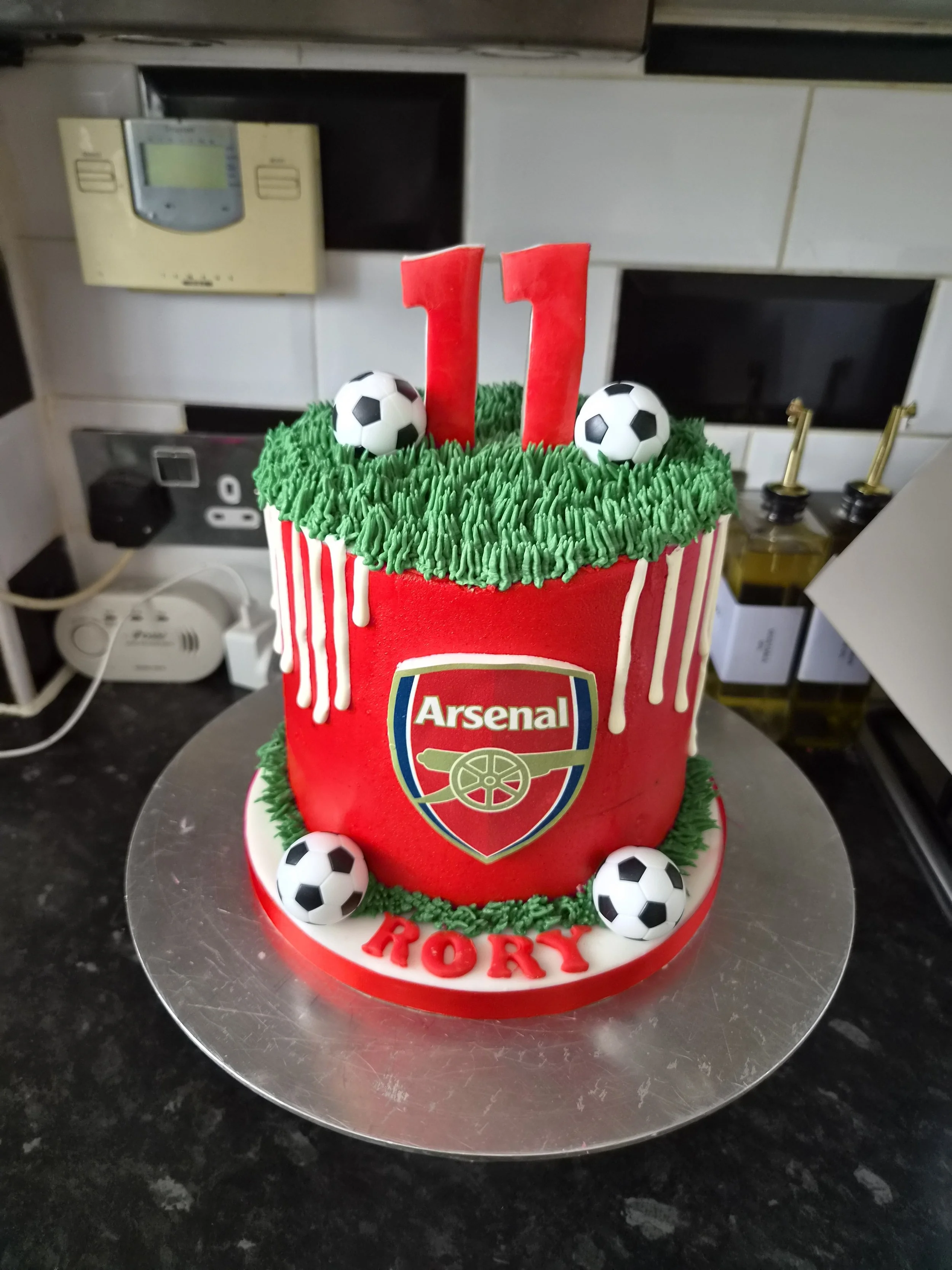 A soccer-themed birthday cake with green icing, soccer ball decorations, and a red number 11 candle on top. The cake features the Arsenal football club logo and the name 'HORY' written on the base. Best birthday cake ever