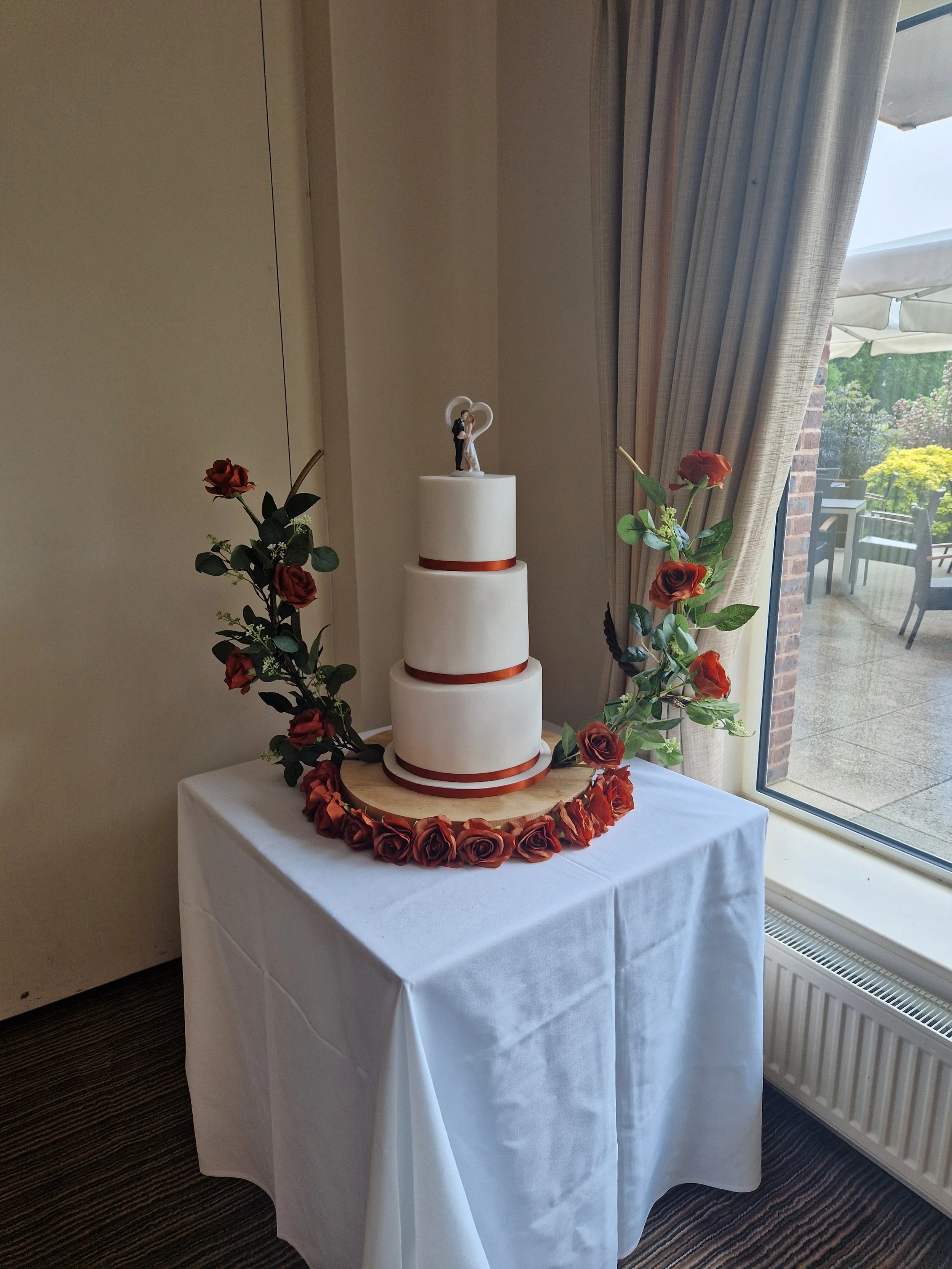 A wedding cake with three tiers, decorated with red ribbons, on a wooden cake stand, surrounded by red and green/artificial flowers, placed on a white tablecloth next to large curtains and a window overlooking an outdoor patio.