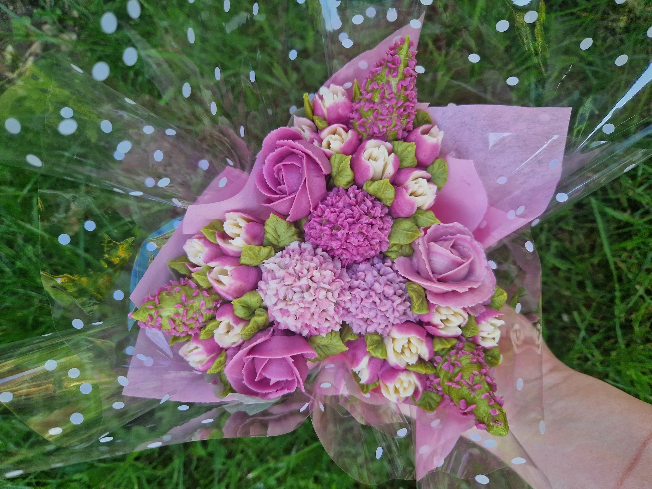A bouquet of pink and white flowers wrapped in pink paper and surrounded by a clear polka-dot cellophane, held against a grassy background.