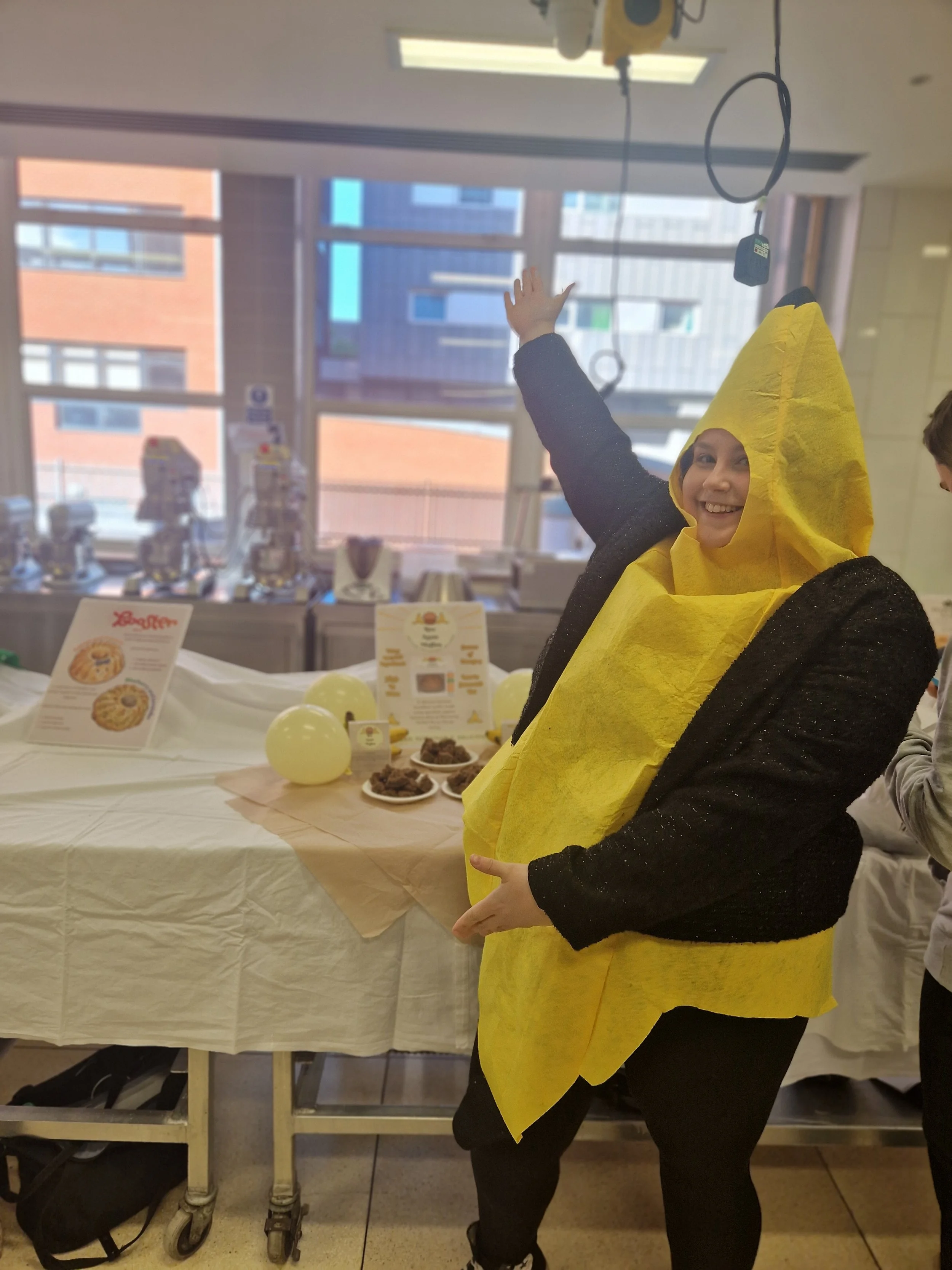 Person wearing a bee costume with a yellow hood and black outfit, smiling and gesturing with an arm extended in a room with large windows and a table displaying promotional materials and yellow balloons.