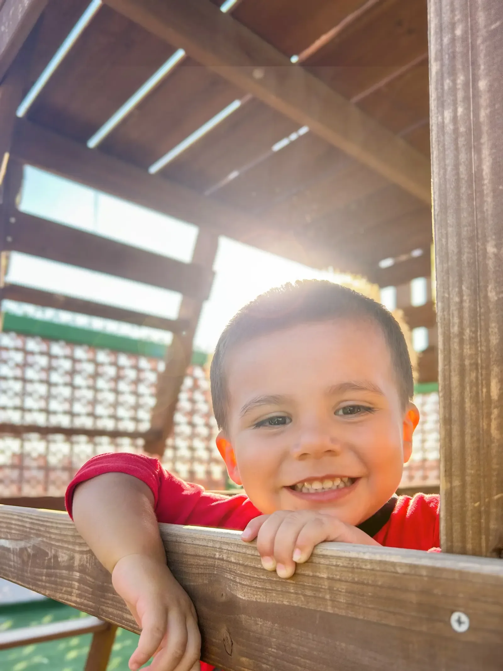 A young Mexican boy smiling with his arms over a wooden rail of playground equipment with the sun shining behind him at St. Innocent Orphanage