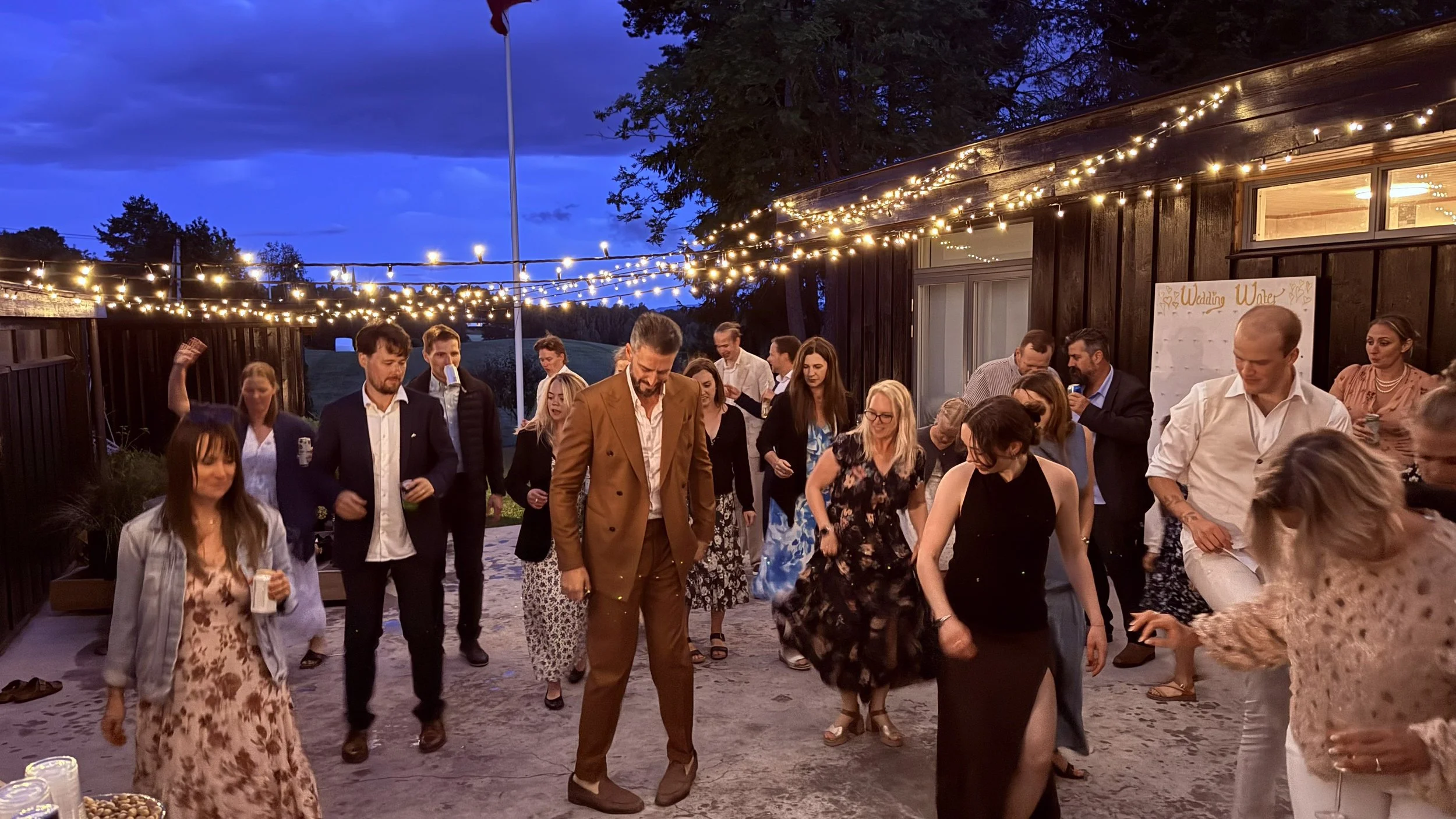 En gruppe mennesker danser på en uteplass under julelys om kvelden, med en mørk bygning i bakgrunnen og en blå kveldshimmel.