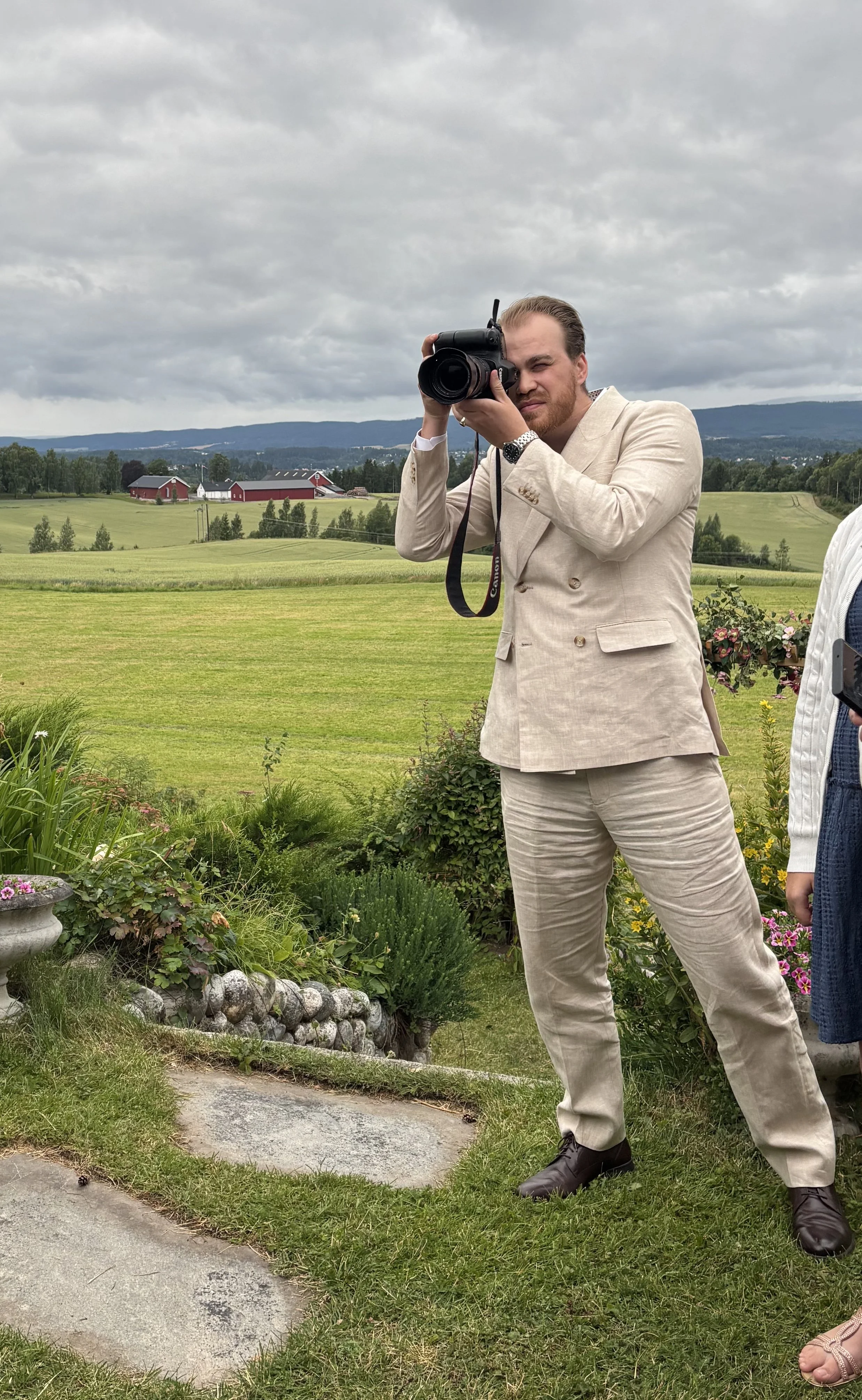 En mann tar et bilde med et digitalt kamera i et landskap med grønne åkrer og en række røde stabbur under en overskyet himmel.