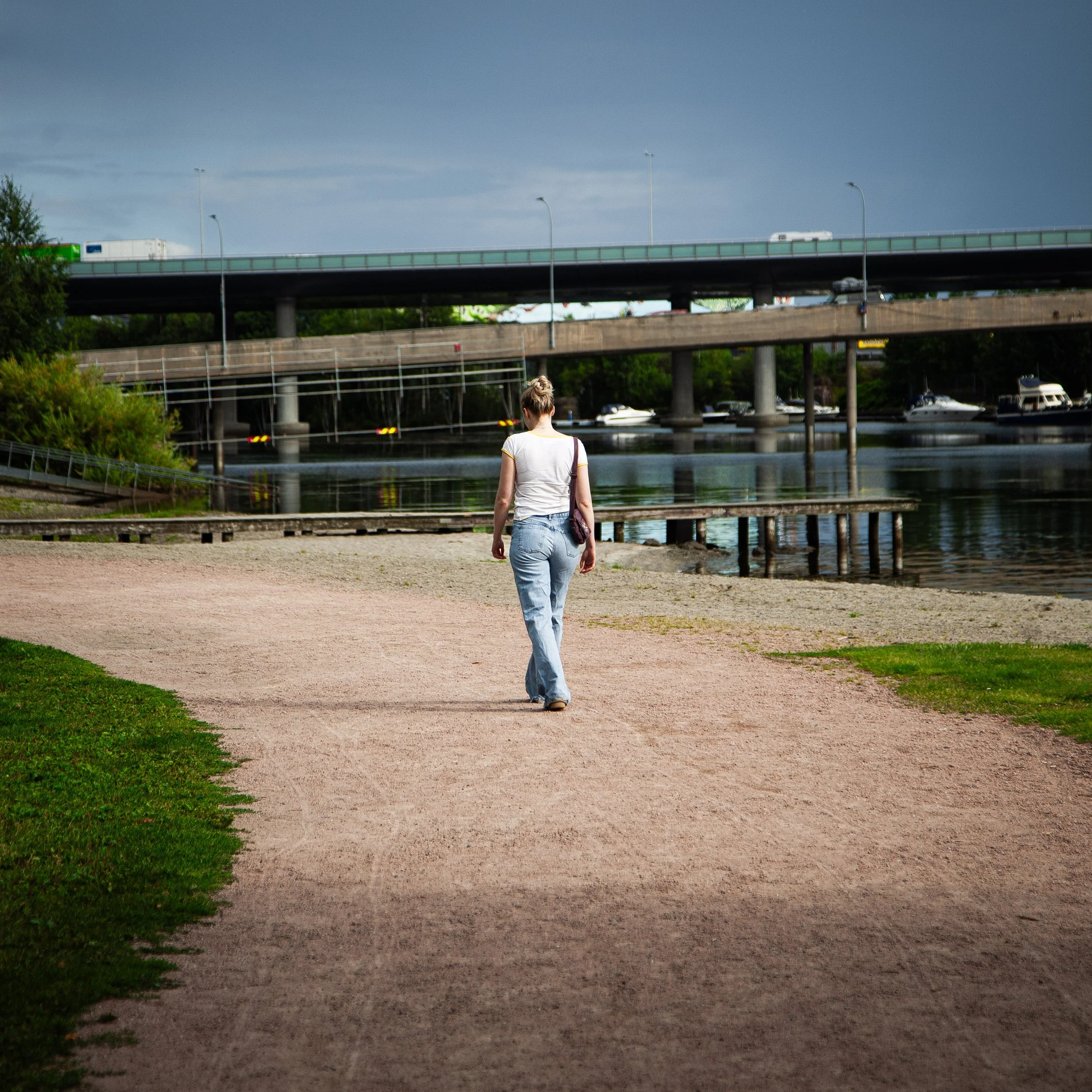 En person går langs en vannkant under en bro med trafikk, med skip på vannet og en by i bakgrunnen.