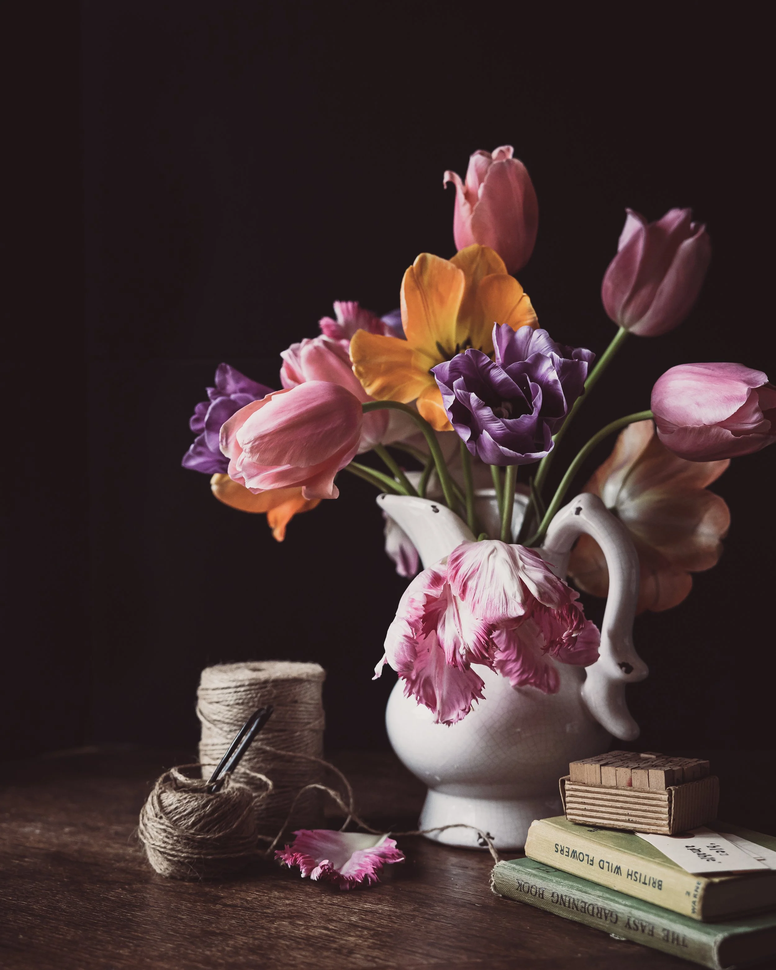 A bouquet of pink, purple, and yellow flowers in a white ceramic vase on a wooden table. There are a few fallen petals on the table, a spool of twine, and three stacked books titled "The Easy Caribbean Cook", "British Wild Flowers", and another book with partly visible text. The background is dark and the lighting is soft.