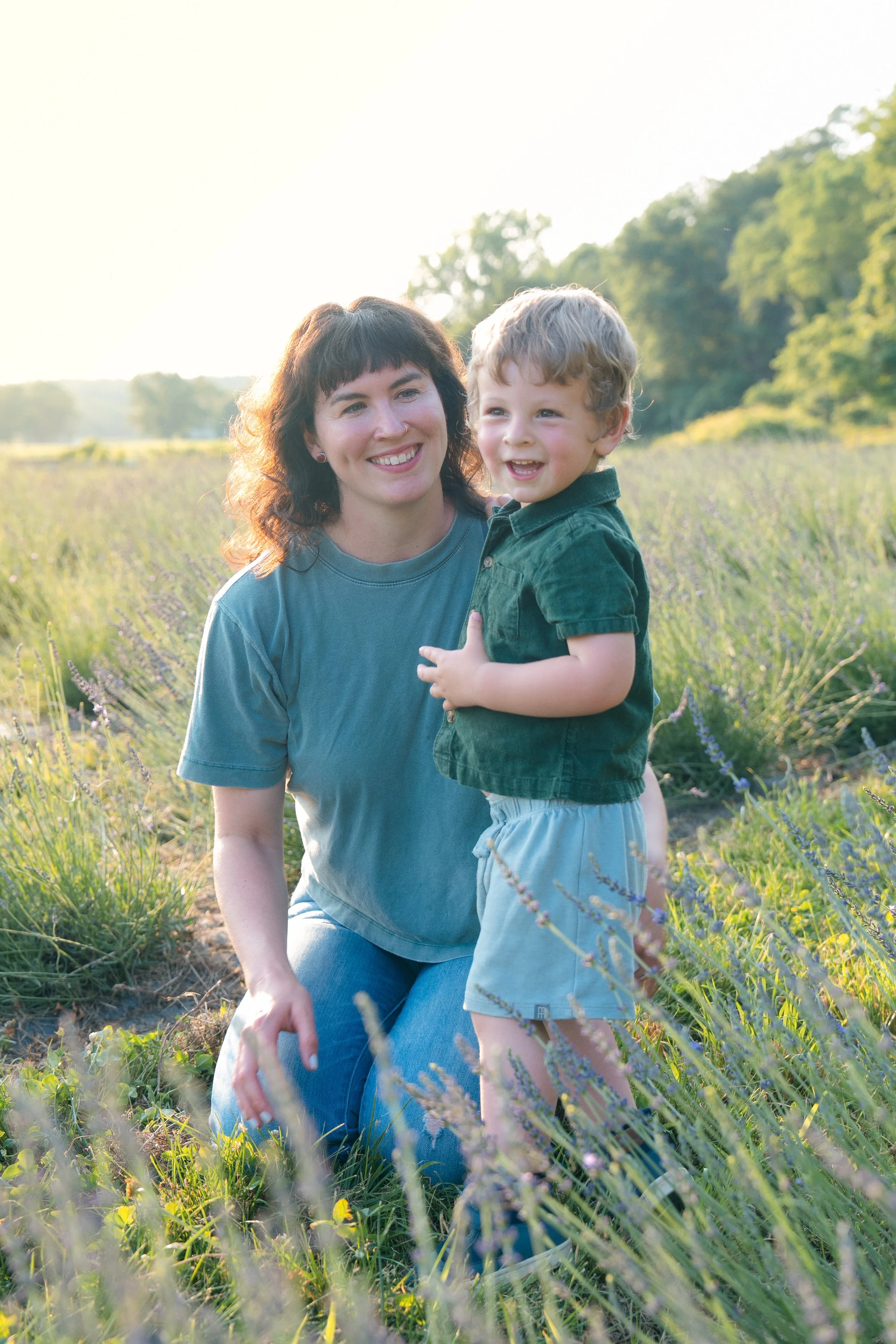A woman and a young boy smiling outdoors in a field of purple flowers with trees and bright sky in the background.