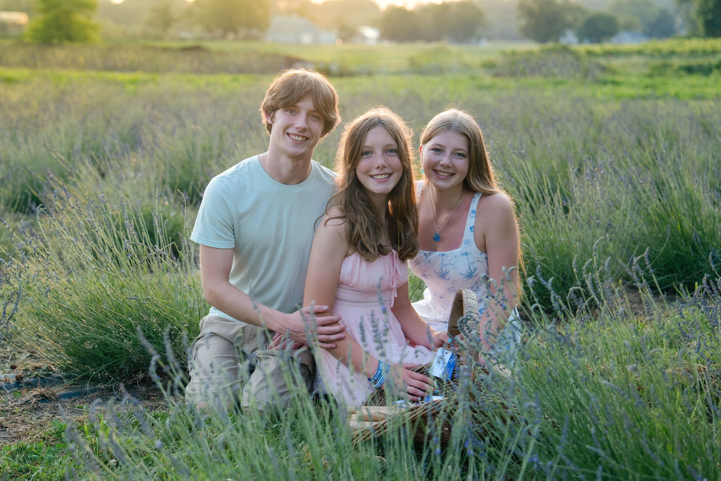 Three young people, one man and two women, sitting in a field of lavender during sunset, smiling at the camera.