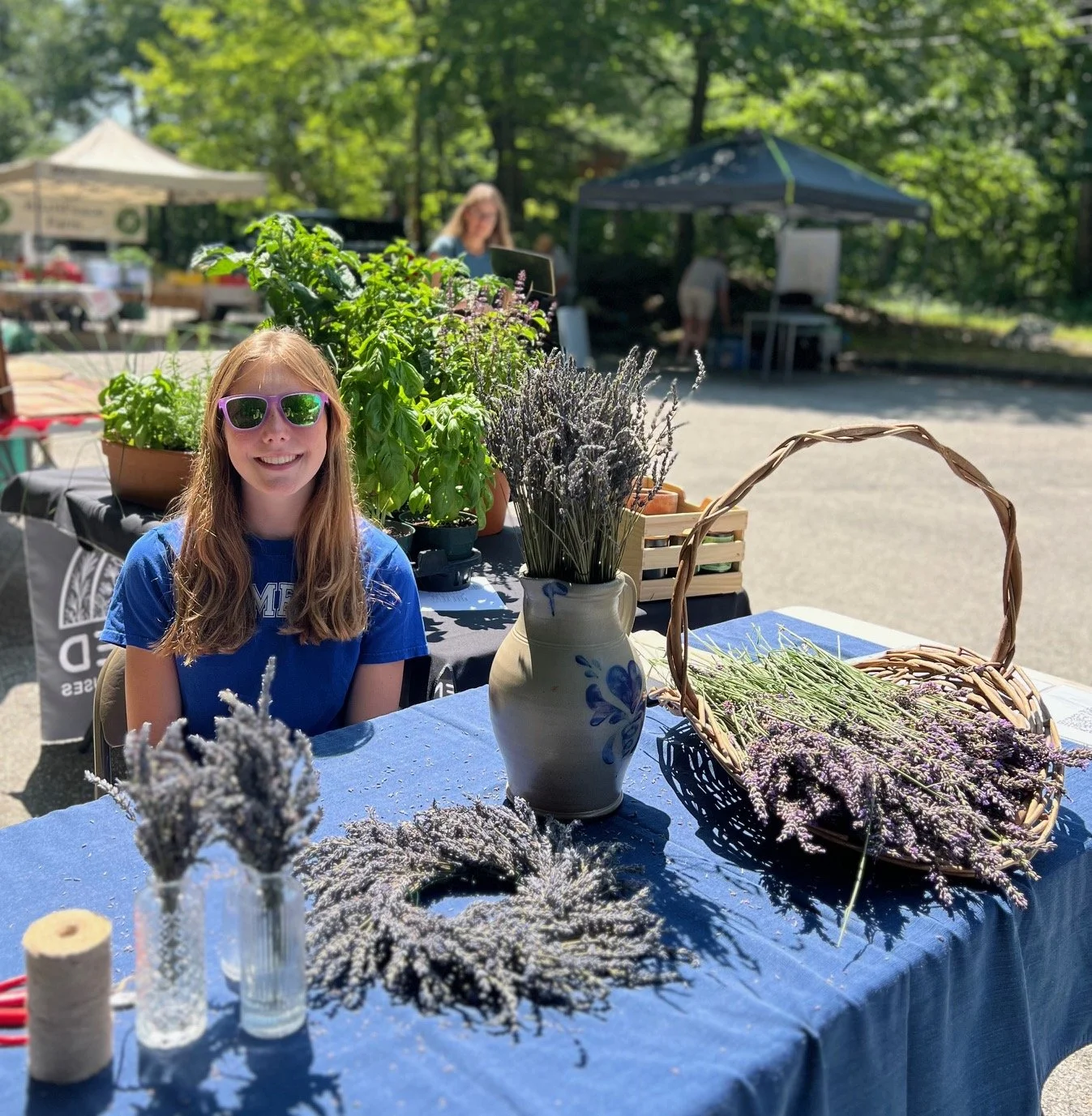A young woman wearing sunglasses and a blue T-shirt sitting at a booth of potted plants and lavender flowers at an outdoor market on a sunny day.