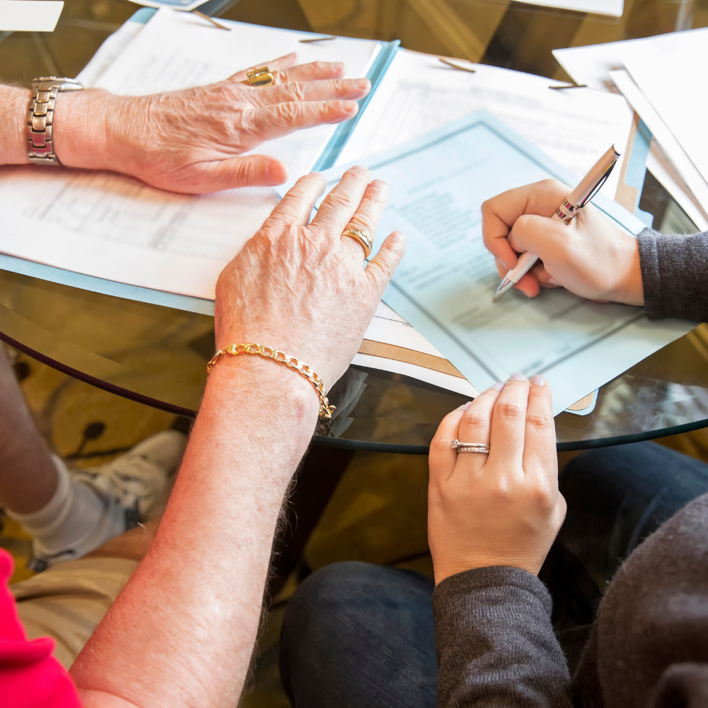 Hands resting on contract pages while another person signs a form at a glass table
