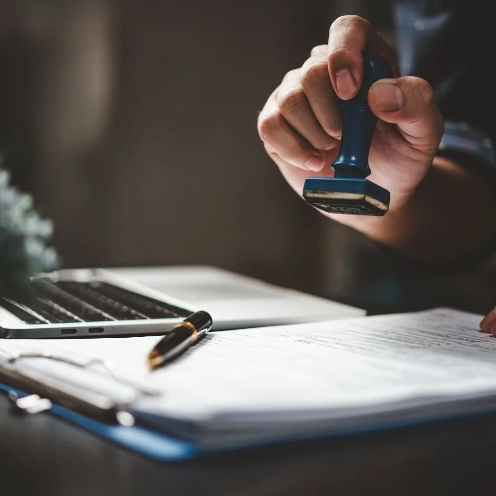 Hand pressing a blue notary stamp over a stack of documents with a pen nearby
