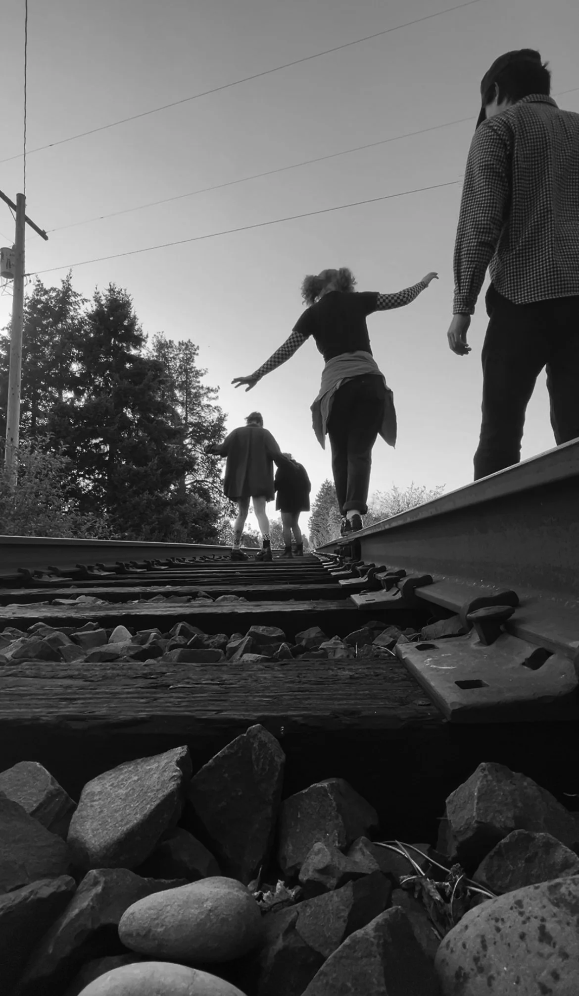People walking along train tracks with trees and power lines in the background, viewed from ground level in black and white.
