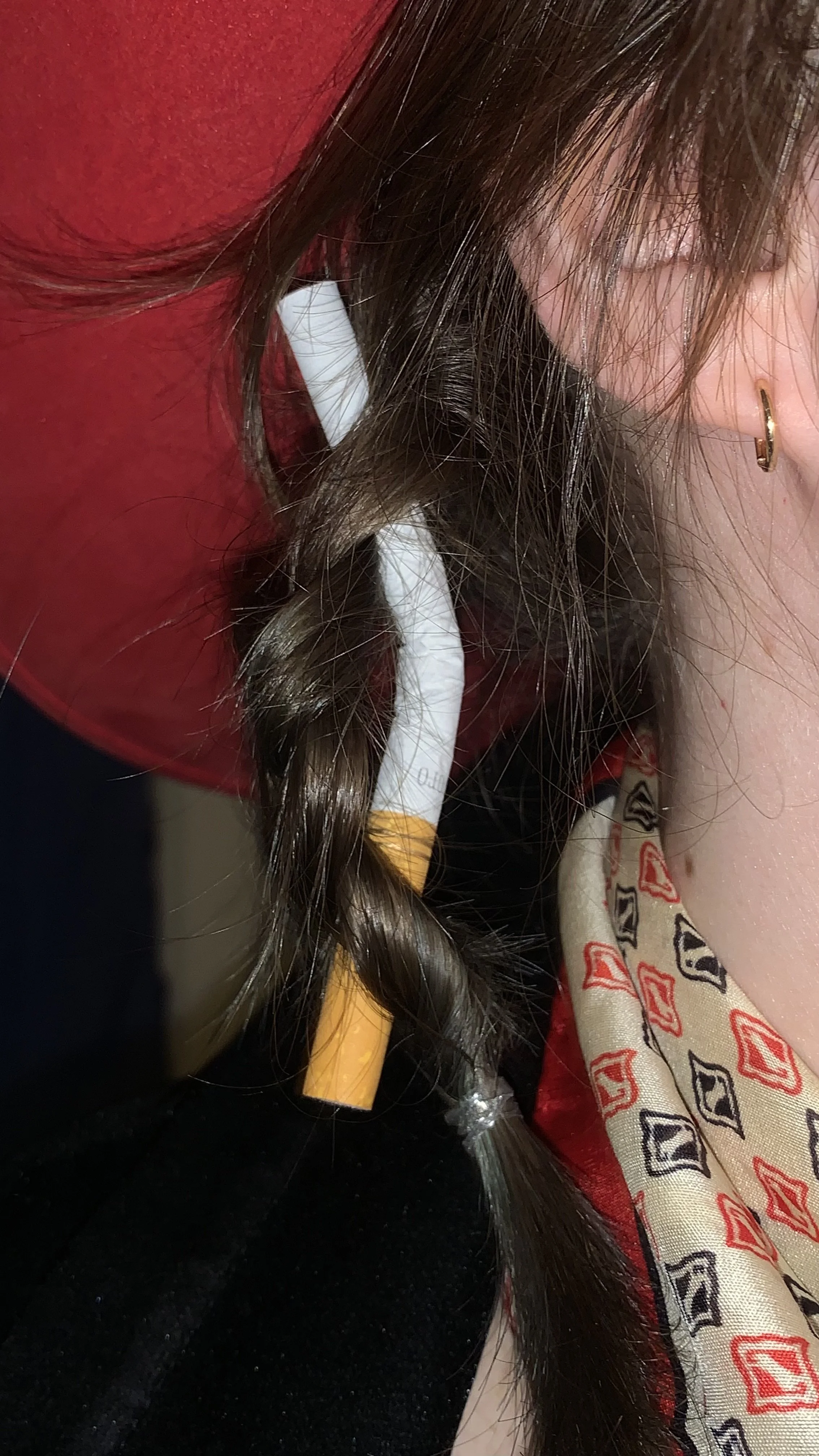 A close-up of a person's ear with a small gold hoop earring, brown hair, a red shirt, a patterned scarf with red and black geometric designs, and a black background.