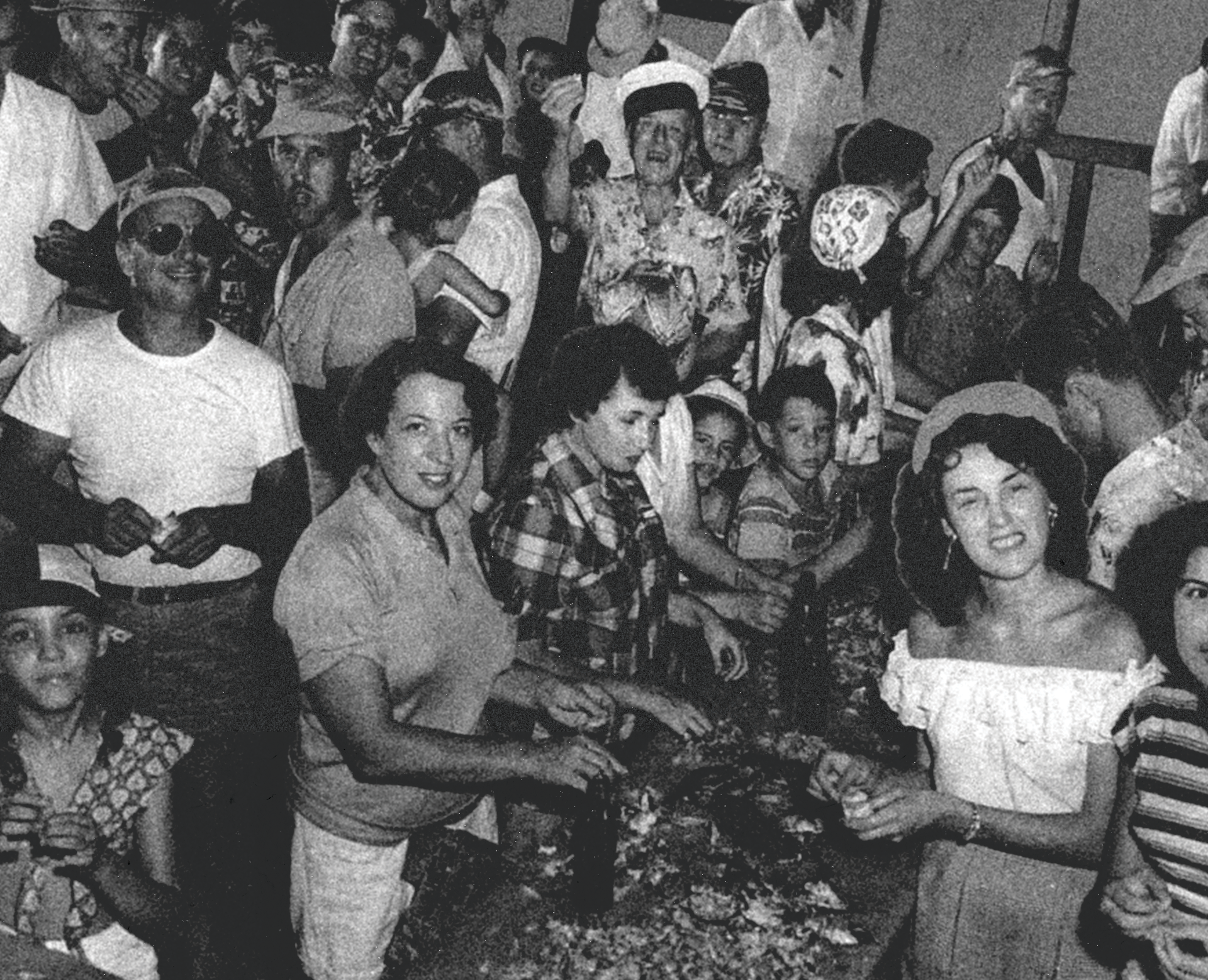 Black and white photo of a group of people, mostly women, gathered at an indoor event, smiling and dressed in casual and traditional clothing eating oysters in Louisiana.