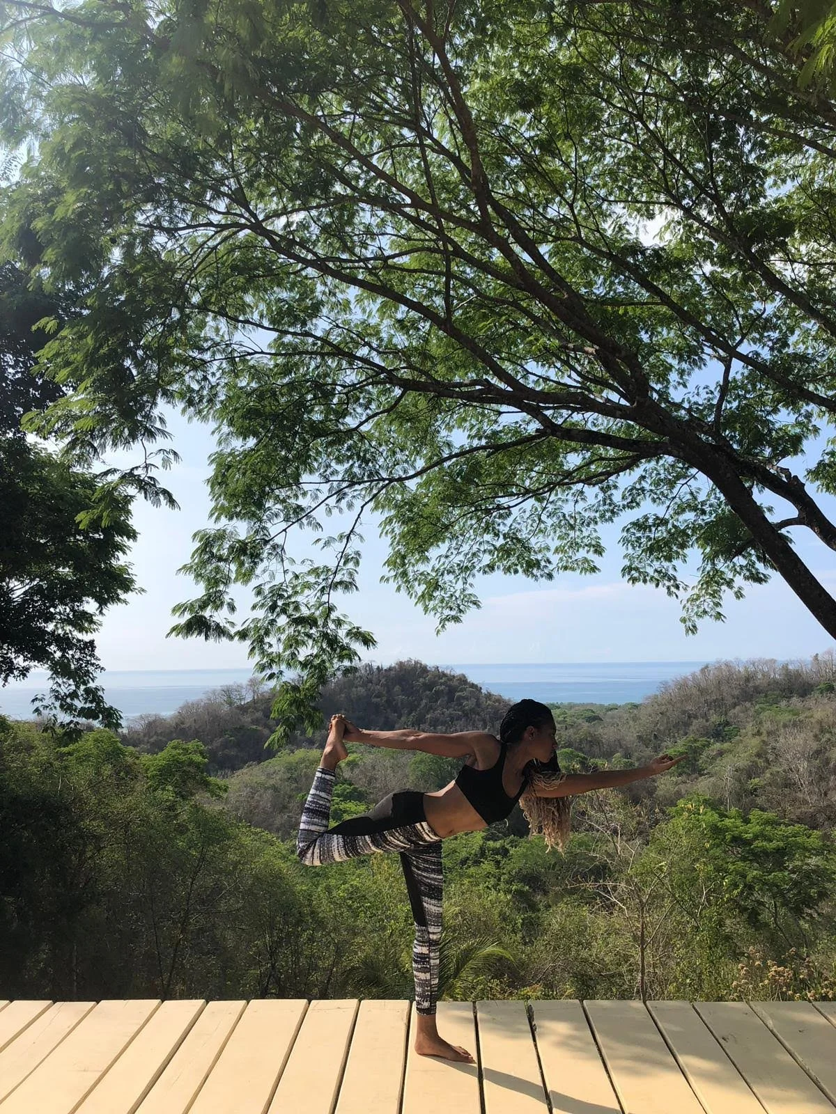 A woman practicing yoga outdoors on a wooden deck, standing on one leg with her other leg bent behind her and foot held by her hand, extending her arm forward in a yoga pose with a scenic view of trees, hills, and the ocean in the background.