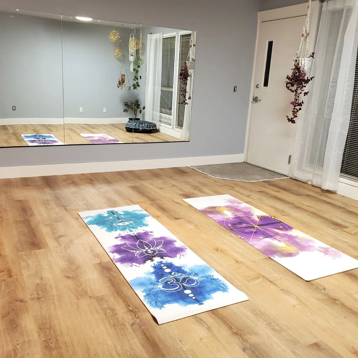 Yoga studio with two mats on wooden floor, a large wall mirror, potted plants, hanging decorations, door with a small mat, and white curtains.