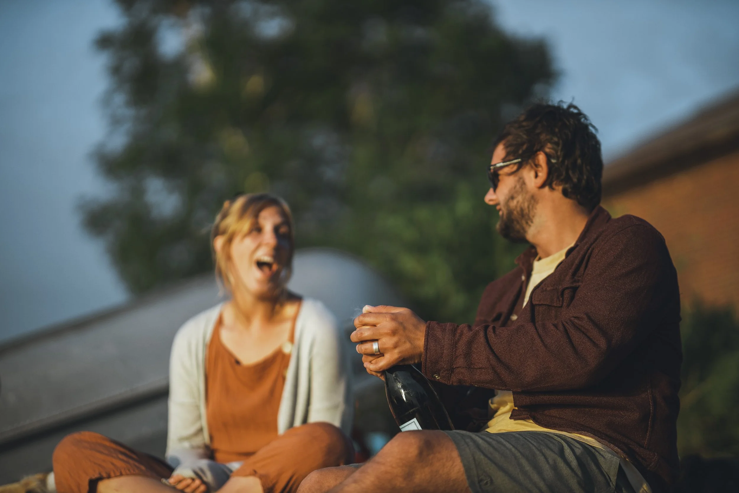 A man with glasses and a woman with blonde hair, both smiling and sitting outdoors at dusk, sharing a moment with a bottle of wine.