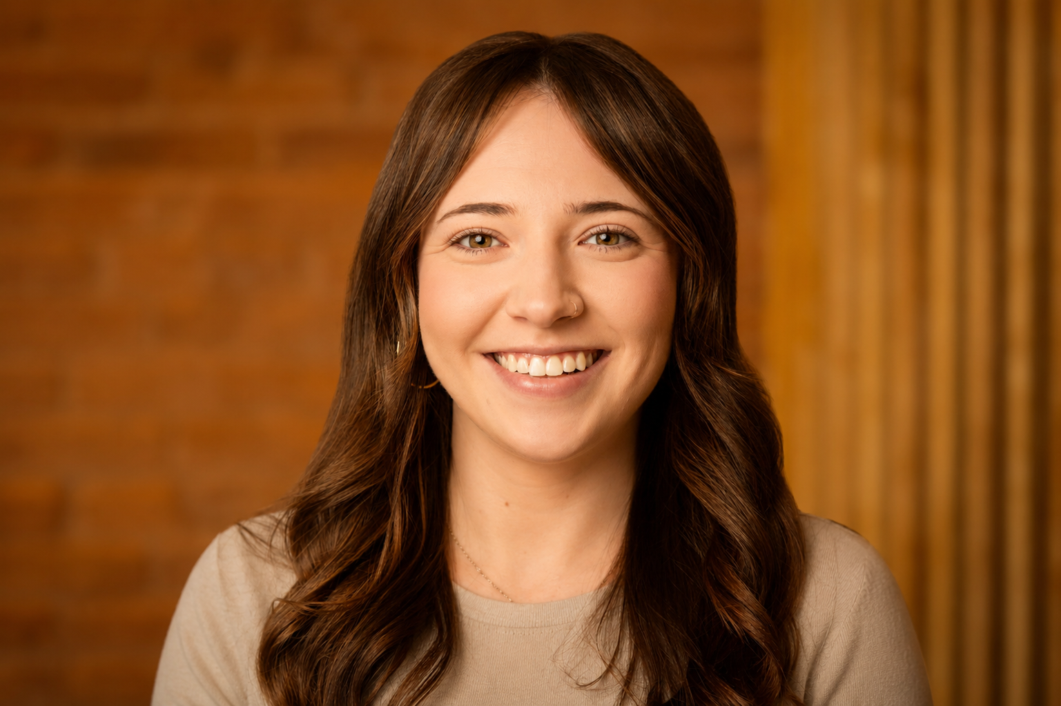 A smiling young woman with long brown hair, wearing a beige top, standing against a wooden background.