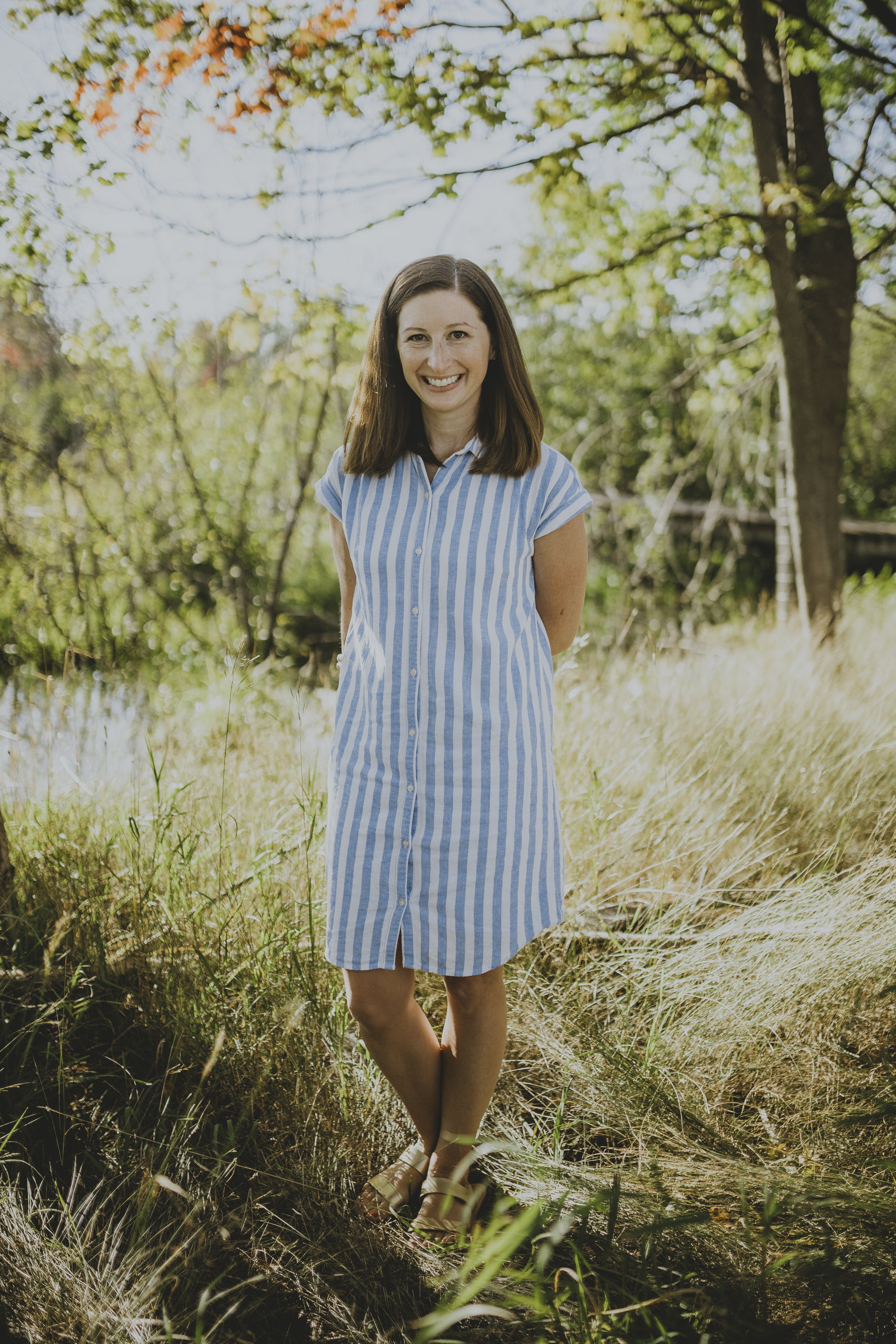 A woman standing outdoors in a grassy area with trees in the background, wearing a blue and white striped dress, smiling at the camera.