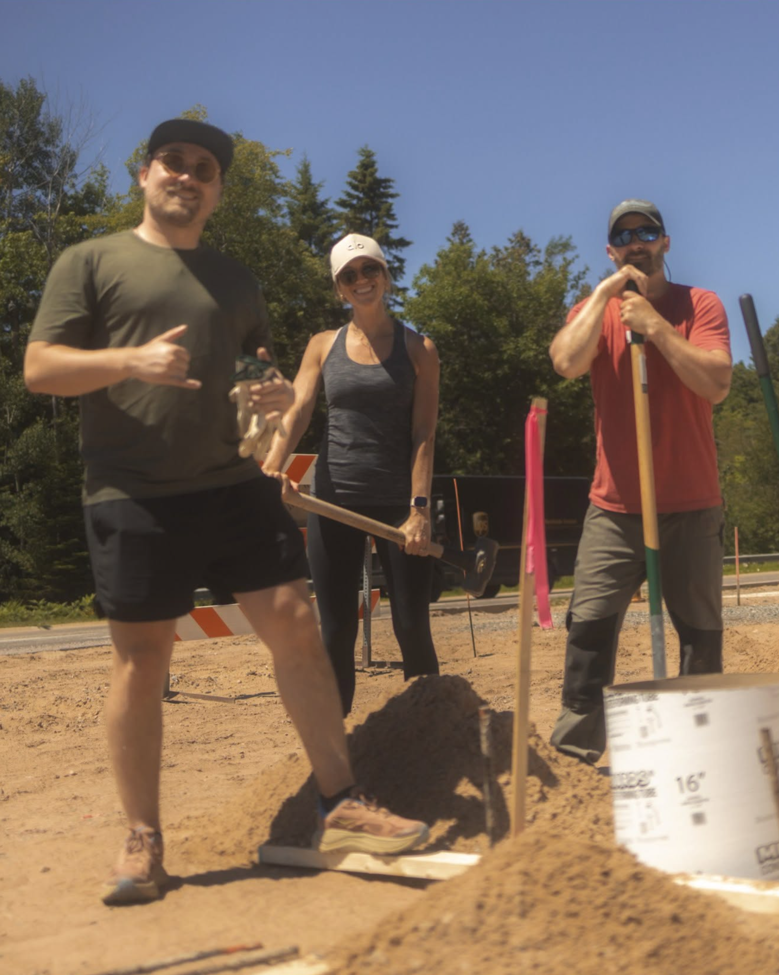 Three people standing at a construction site with trees and blue sky in the background. One person is giving a thumbs-up, another is smiling and holding a tool, and the third is holding a tool with both hands. There are construction materials and items in the foreground.