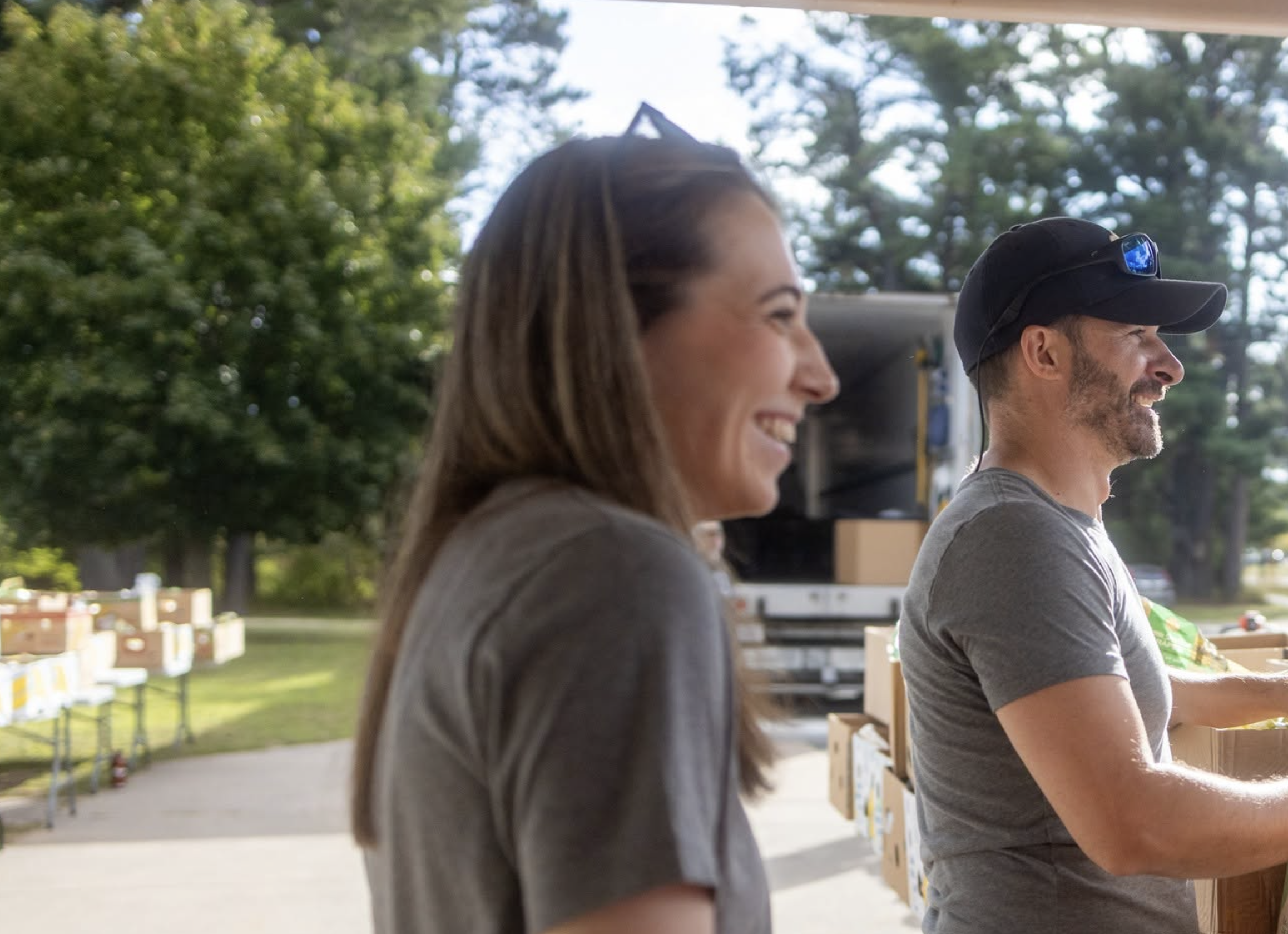 Two smiling people, a woman with long brown hair and a man with a beard, standing side by side at an outdoor event with trees and boxes around, possibly at a donation or distribution site.