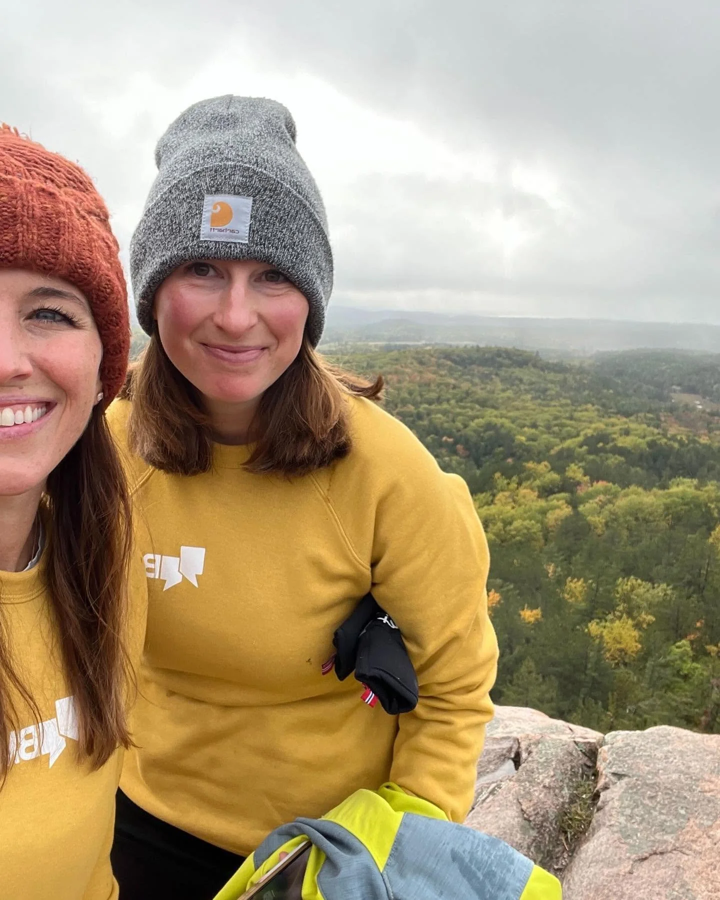 Two smiling women wearing yellow shirts and gray knit hats standing on a rocky overlook with a view of trees and rolling hills in the distance, overcast sky.