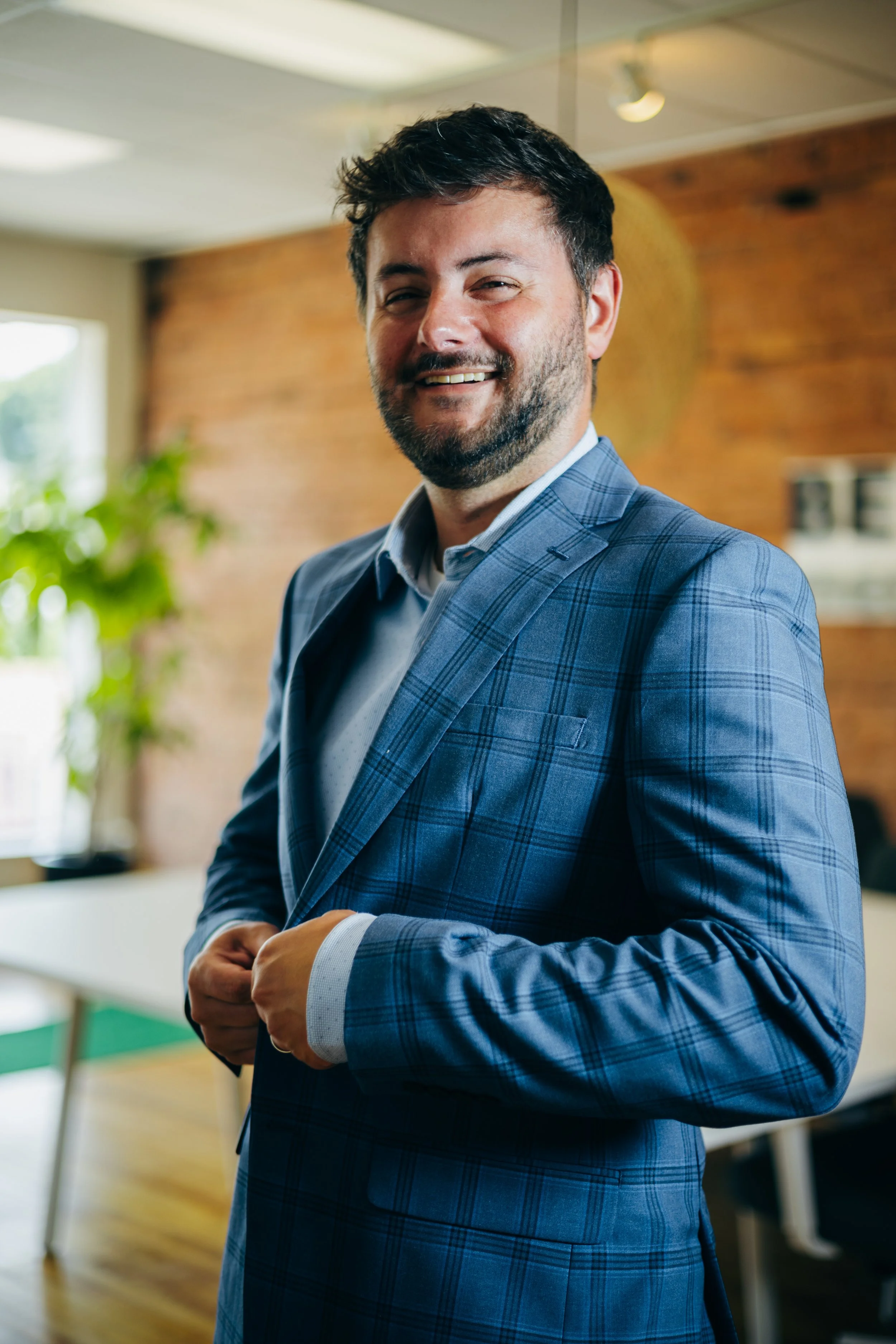 A man with dark hair and a beard smiling, wearing a blue plaid suit, standing indoors with a brick wall and a window in the background.