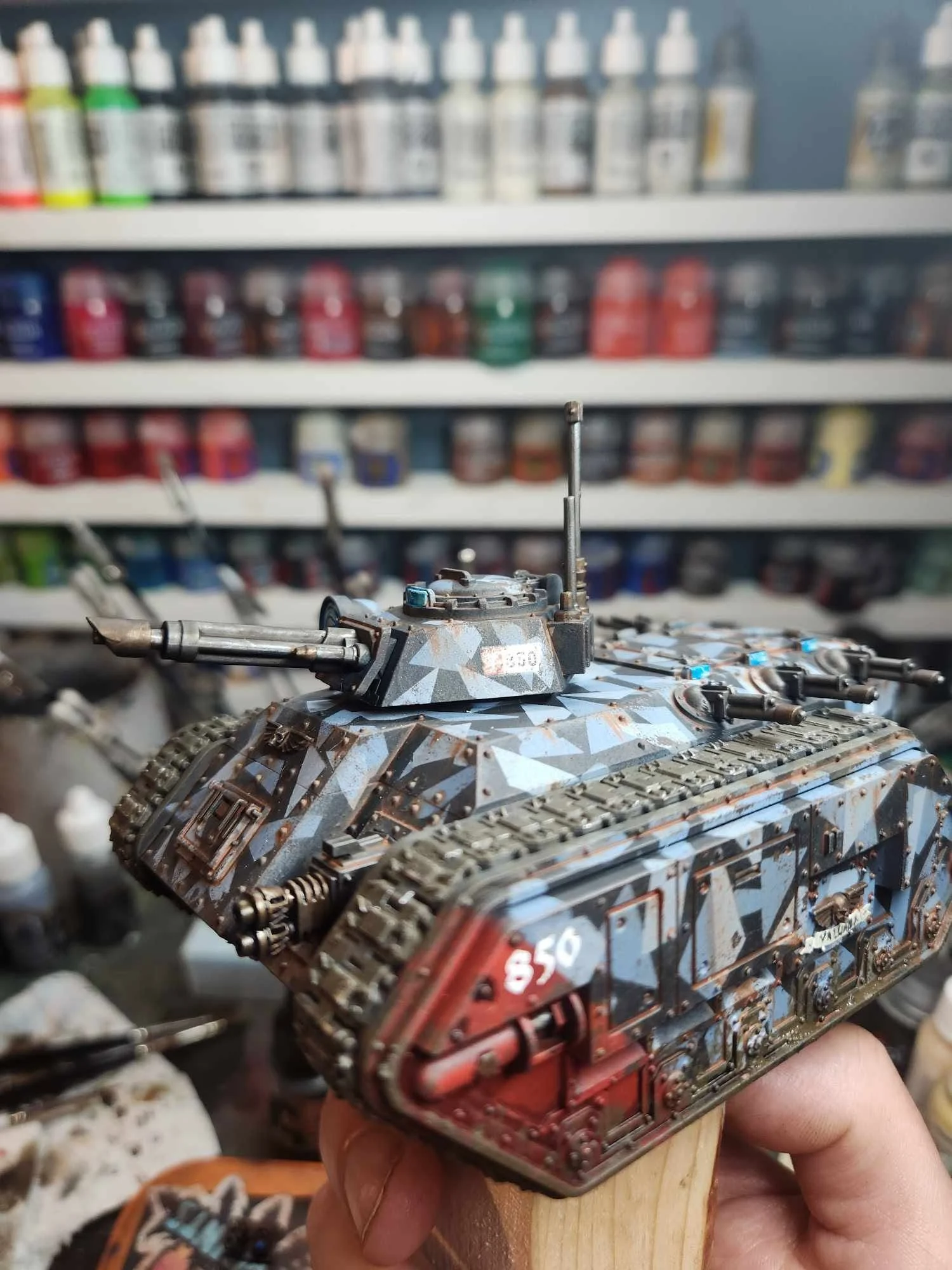 A person holding a detailed, painted miniature model of a tank with a camouflage pattern, in front of shelves filled with paint bottles.