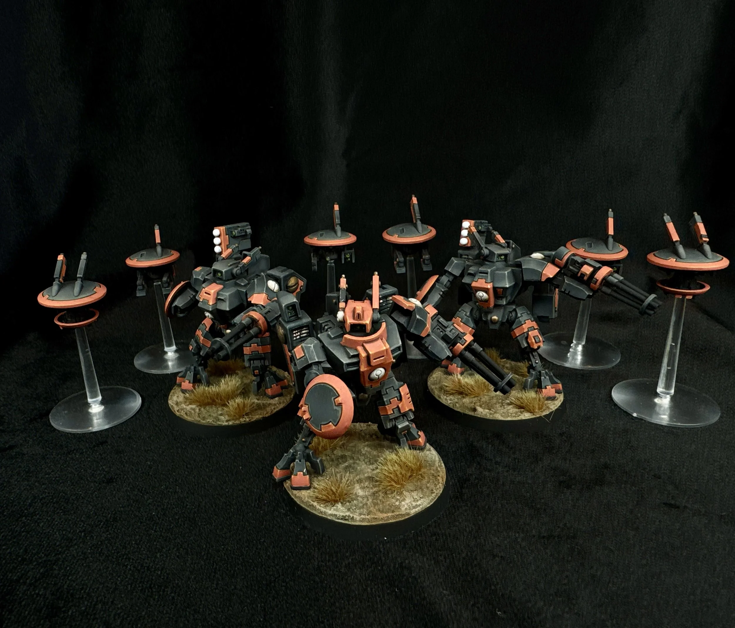 A set of miniature mechs with black and orange armor standing on bases with grass and dirt textures, displayed on a black surface with a black background.