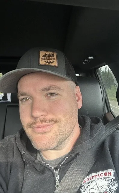 A man with a cap and hoodie sitting in a vehicle, smiling at the camera.
