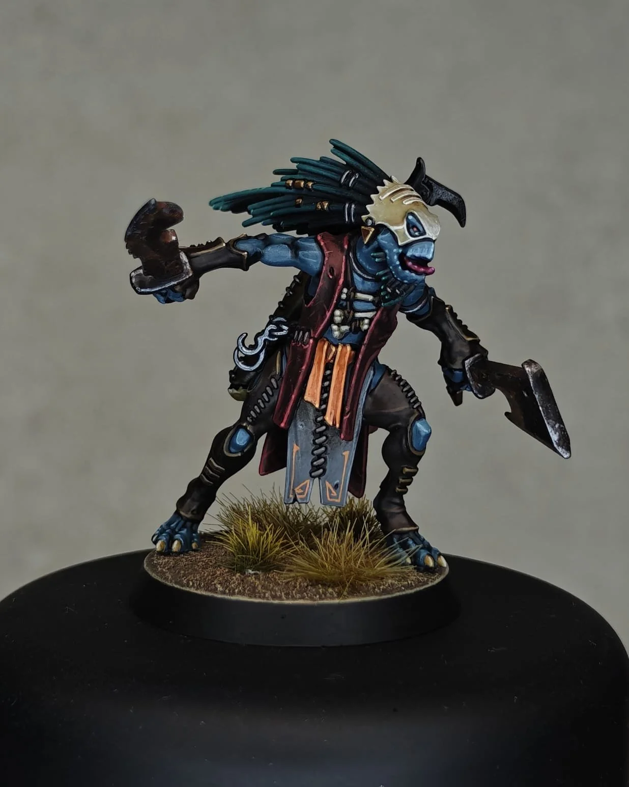 Miniature figurine of an anthropomorphic creature with blue skin, black hair, and a skull mask, dressed in tribal clothing, standing on a round base with grass tufts, holding bladed weapons in both hands.