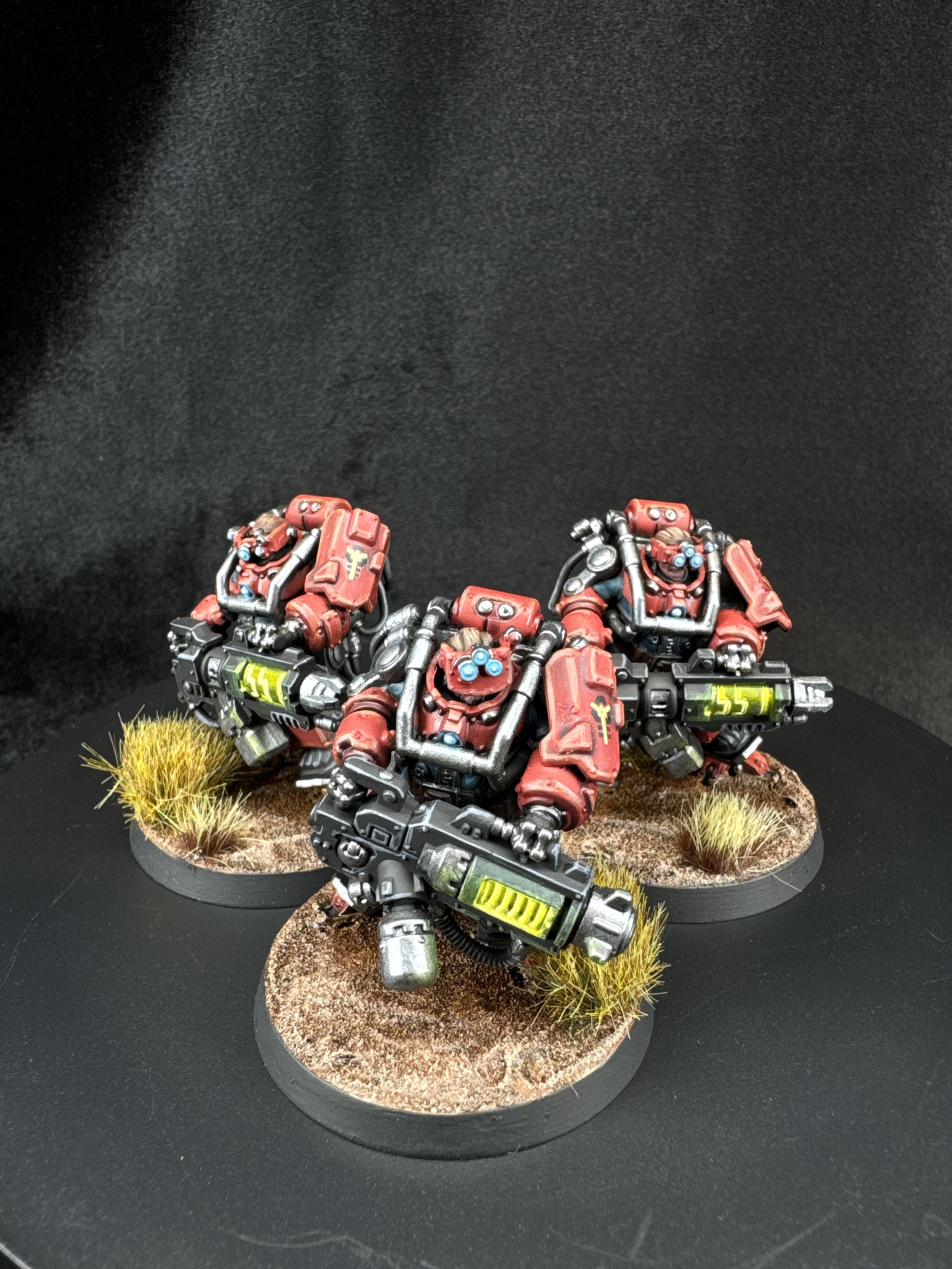 Three miniature models of red and black futuristic soldiers with guns, standing on round bases with textured terrain and grass tufts, against a black background.