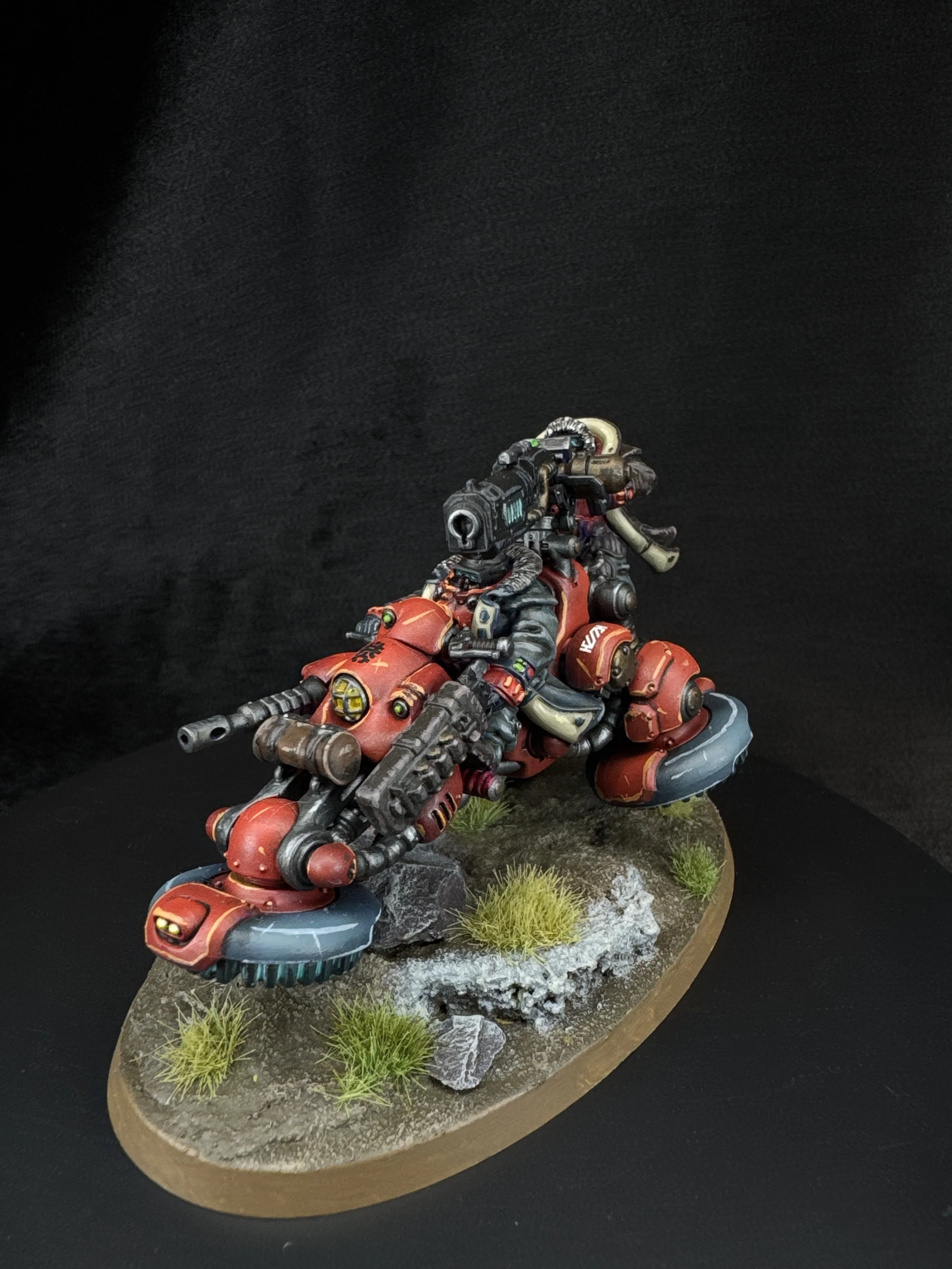 A detailed miniature model of a futuristic red motorcycle with advanced technology features, mounted on a textured base with grass and rocks, against a black background.