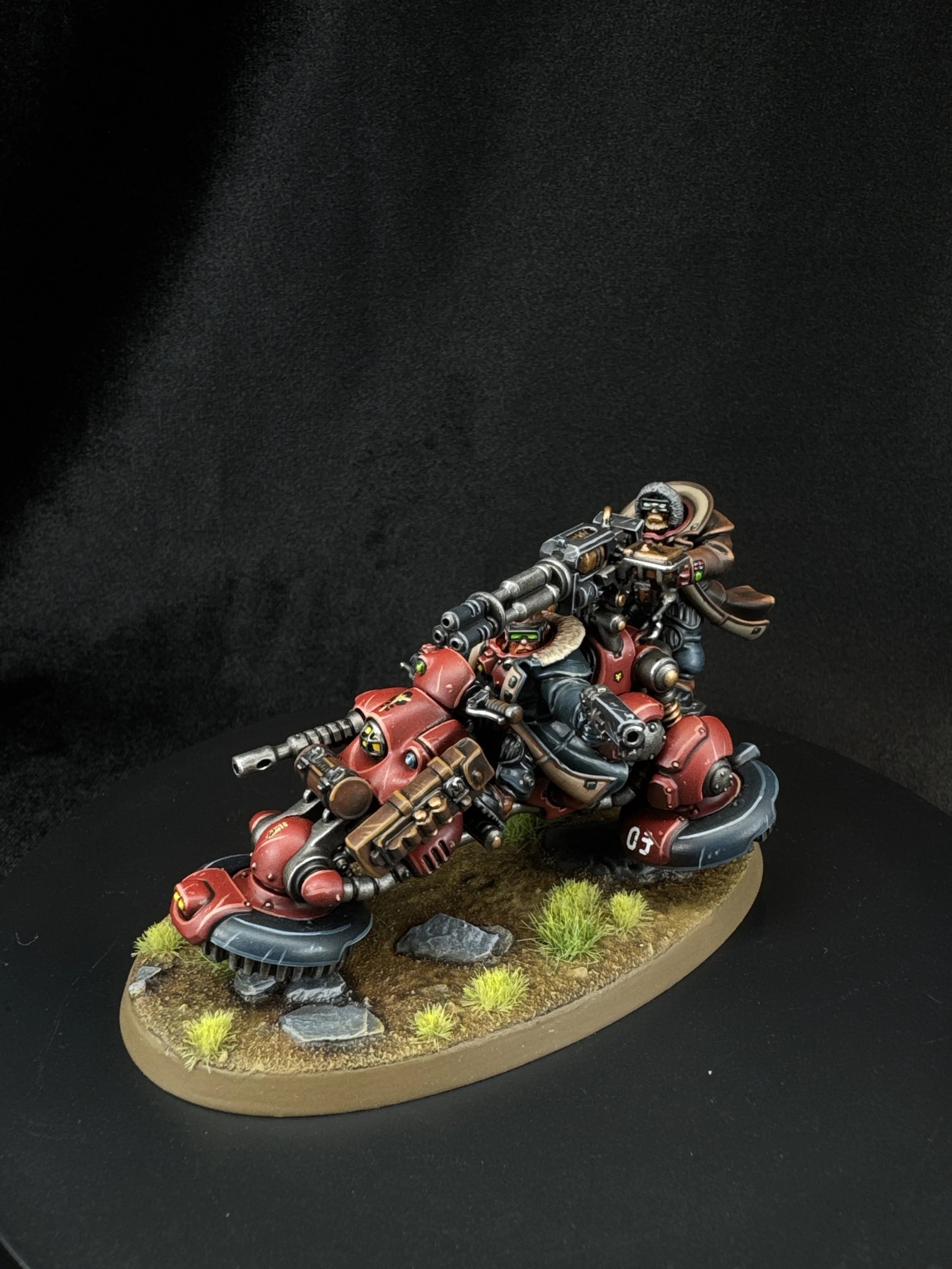 Miniature model of a futuristic red motorcycle with two riders, set on a grassy base with rocks and small plants, against a black background.