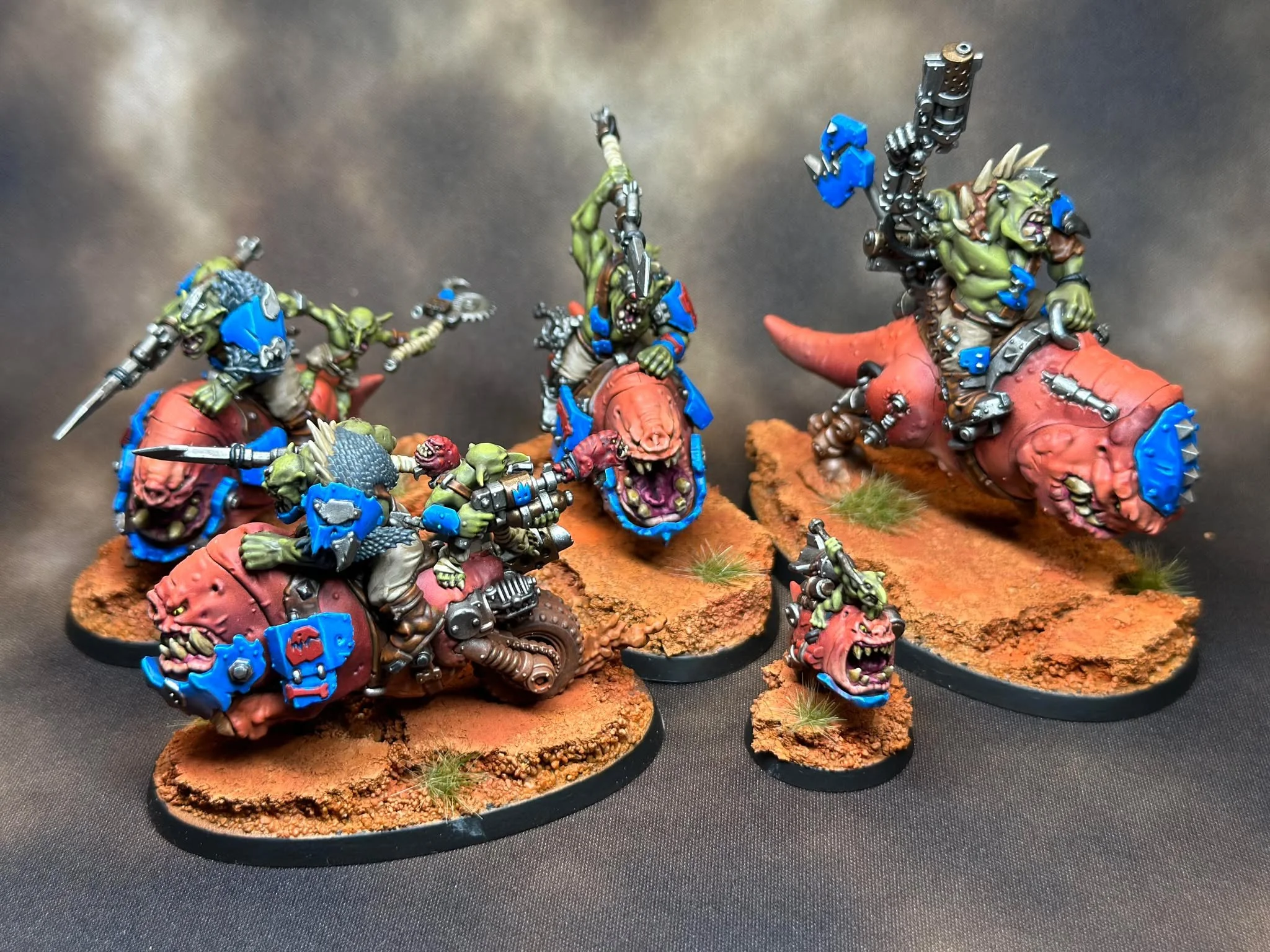 Miniature tabletop figures of orcs riding pig-like creatures, painted in green, blue, and metallic colors, on a textured orange base.