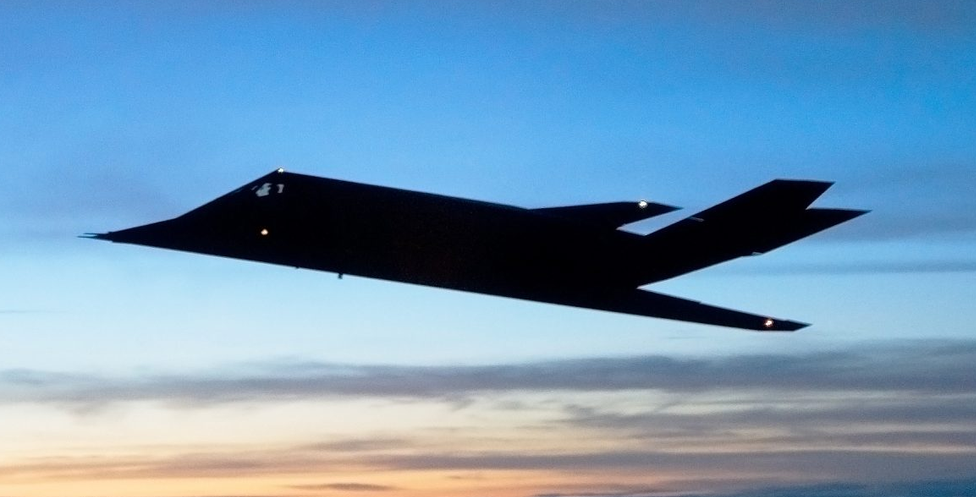 A black stealth bomber aircraft flying in the sky during sunset with a gradient of colors from orange to blue.