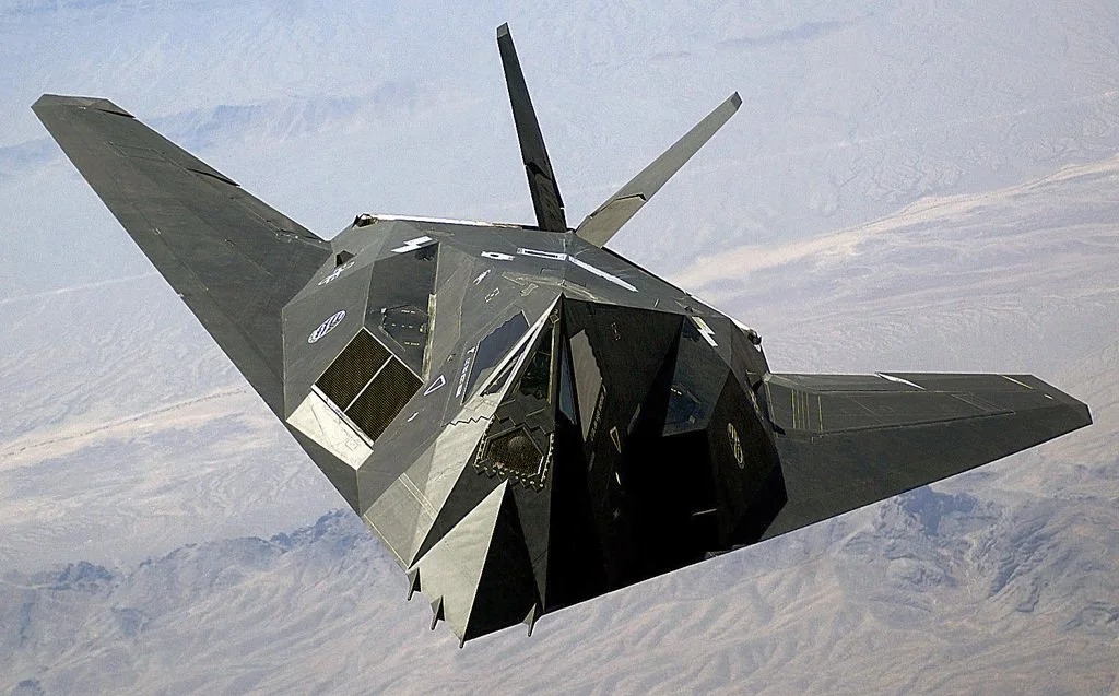 Black stealth fighter jet flying above desert terrain.