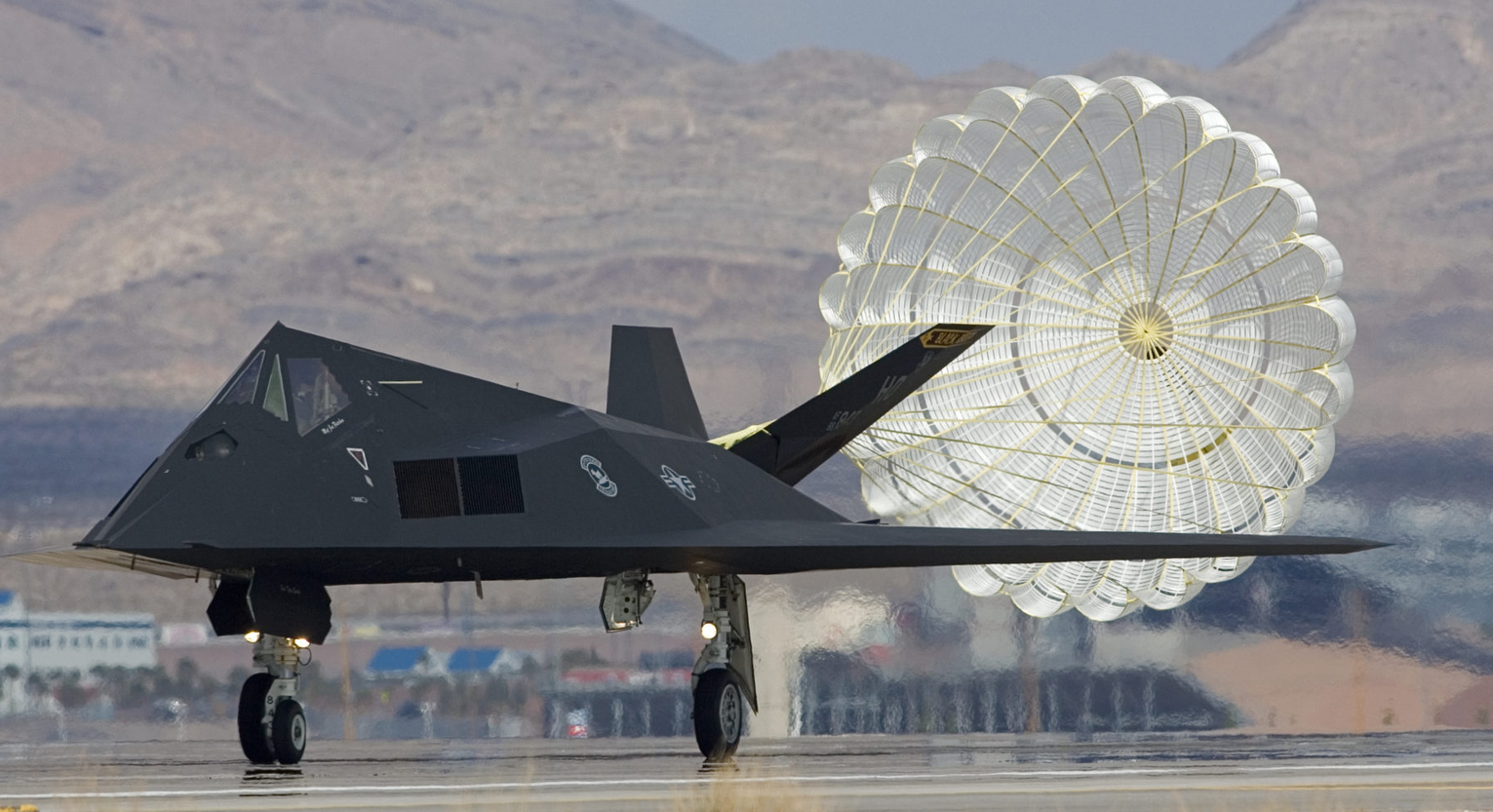 A stealth aircraft on a runway with a large parachute deployed behind it, set against a mountainous background.