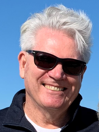 Smiling man with gray hair wearing sunglasses and a dark jacket outdoors against a blue sky.