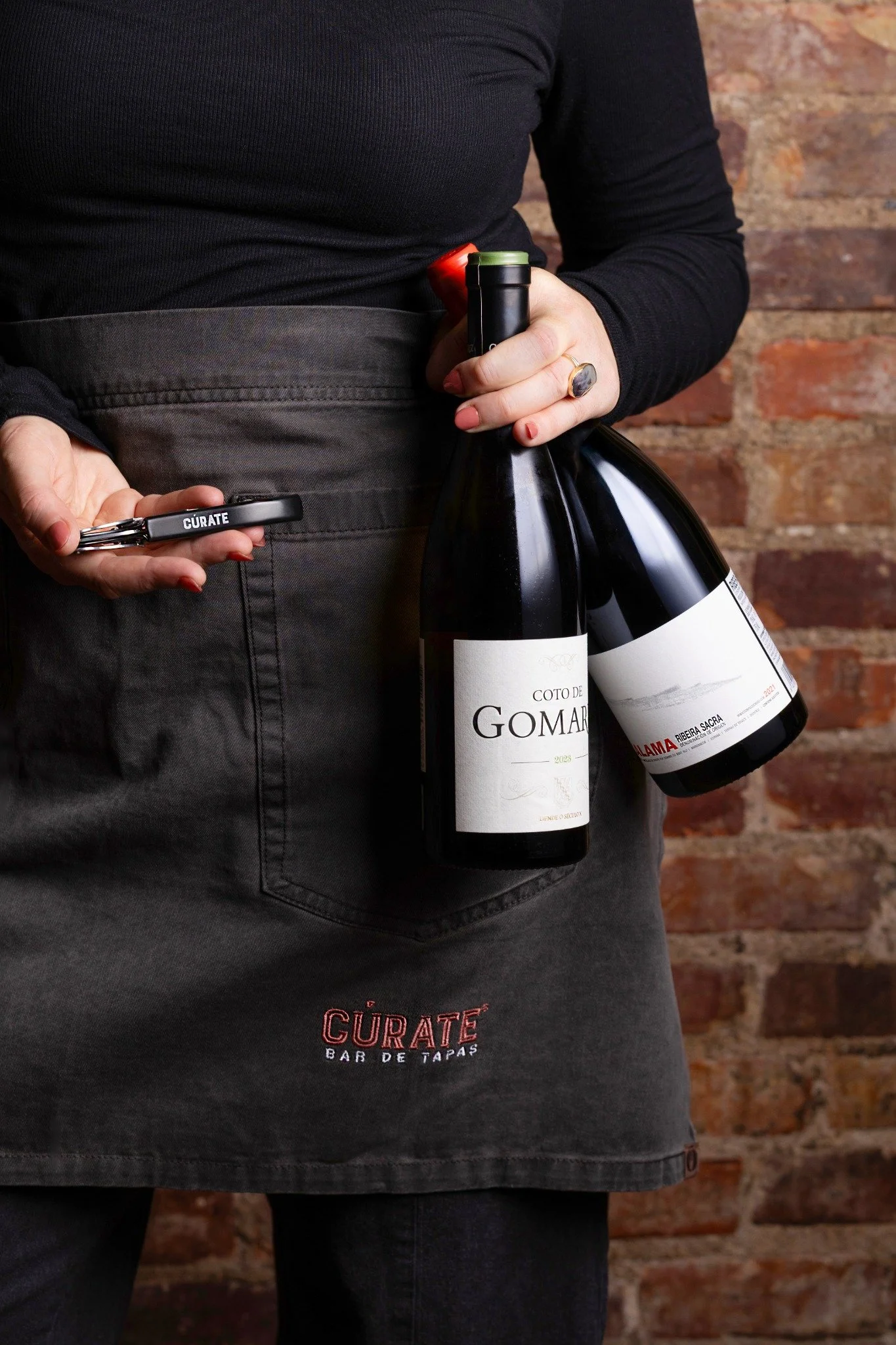 Hospitality — Cúrate wine service with two Spanish bottles and a branded wine key against an exposed brick interior.