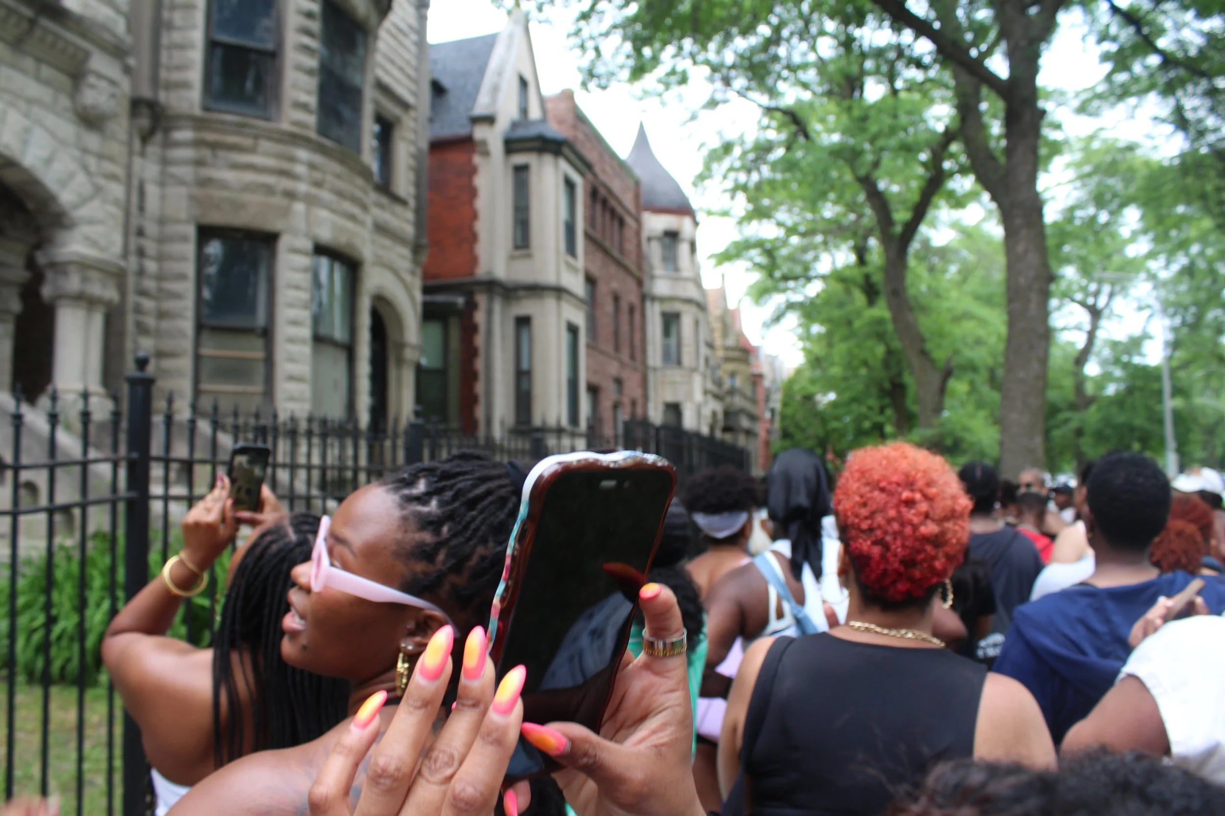 A group of people gathered outdoors near historic buildings with green trees and bright sky in the background. Some are taking photos or selfies, and colorful umbrellas are seen among the crowd.