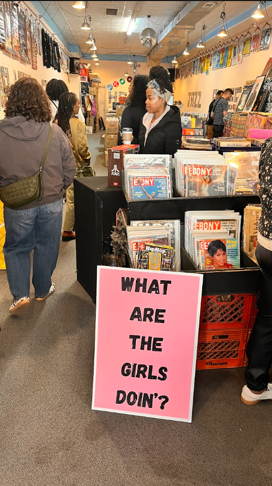 People shopping at a record store, with a pink sign in the foreground that reads "WHAT ARE THE GIRLS DOIN'?" The store has vinyl records, posters, and magazines on display.